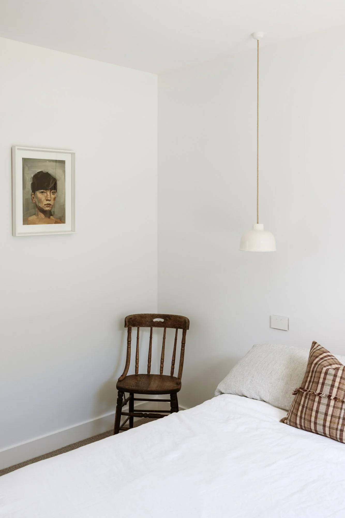 A corner of a bedroom with a white bed, two pillows, a wooden chair, a framed portrait on the wall, and a hanging white lamp.