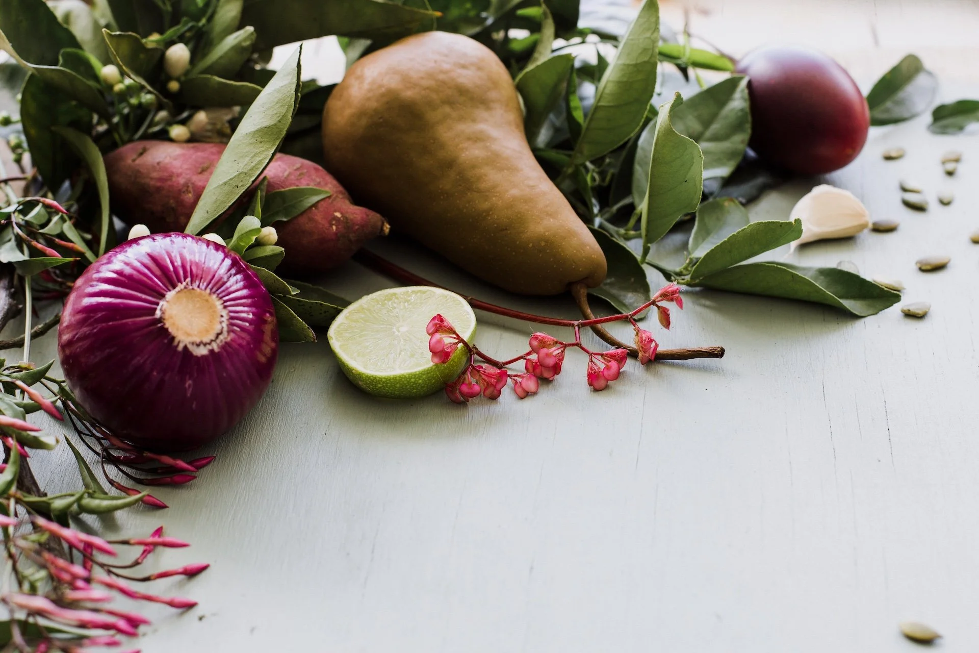 An arrangement of fresh fruits, vegetables, and herbs on a white surface, including a lime half, red onion, a pear, a red potato, a garlic clove, and leafy greens with pink flowers. Photographed for a cookbook cover in Tauranga.