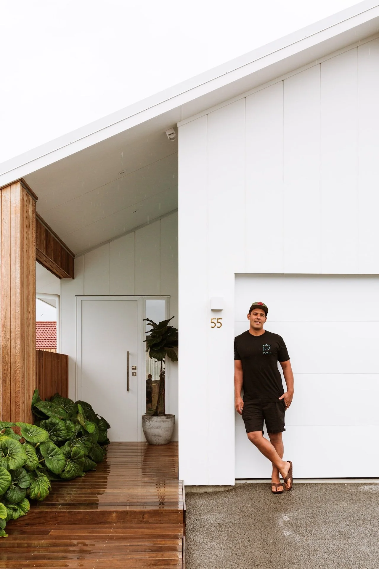 A man standing outside a modern house leaning against a white wall, wearing a black t-shirt, shorts, flip-flops, and a cap, with a house number 55 visible and some green plants nearby.
