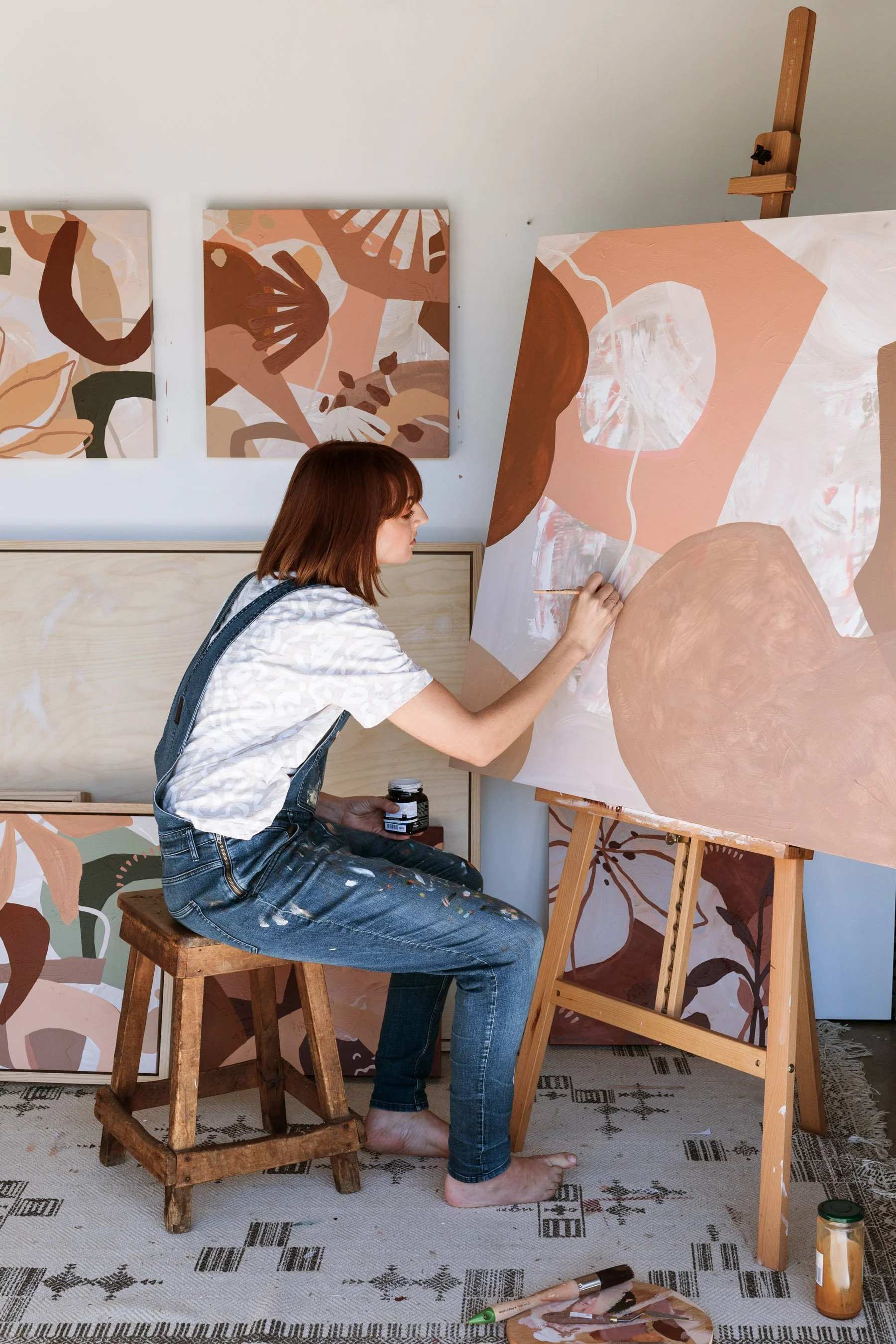 A woman painting abstract artwork on a large canvas in an art studio, surrounded by finished paintings, with a jar of black paint, and painting tools on the floor. Lifestyle commercial branding photography in Mount Maunganui.
