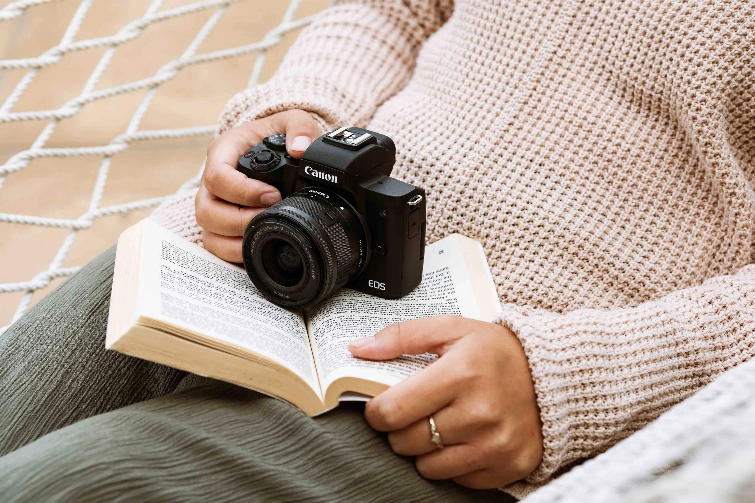 Person sitting on a white netted surface holding a Canon EOS camera with a lens, and reading a book. Photography for an advertising campaign.