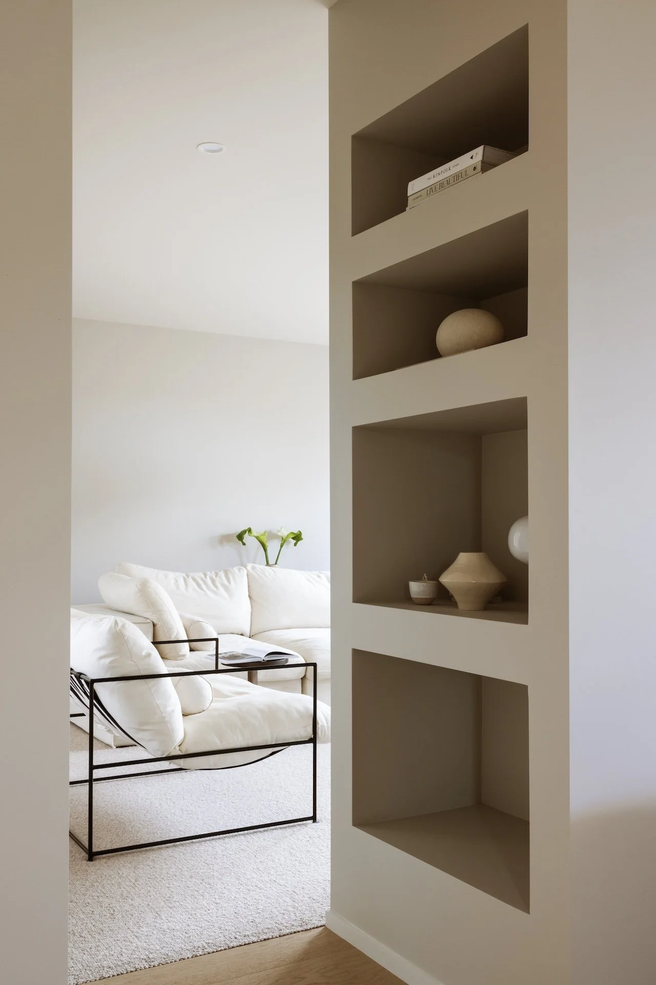 A view of a living room with a white sofa and a black metal armrest, and built-in wall shelves with decorative items.