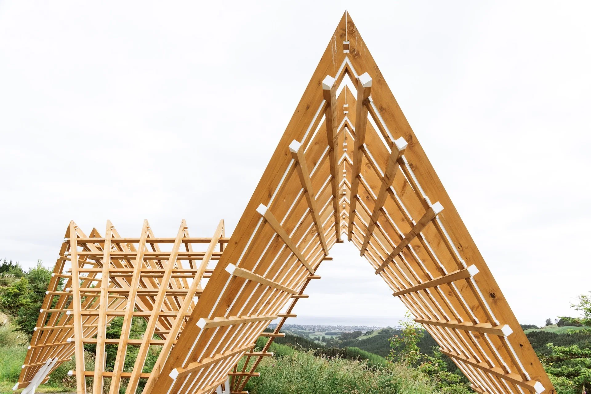 Wooden roof framework architectural sculpture in an outdoor setting with green landscape and cloudy sky.