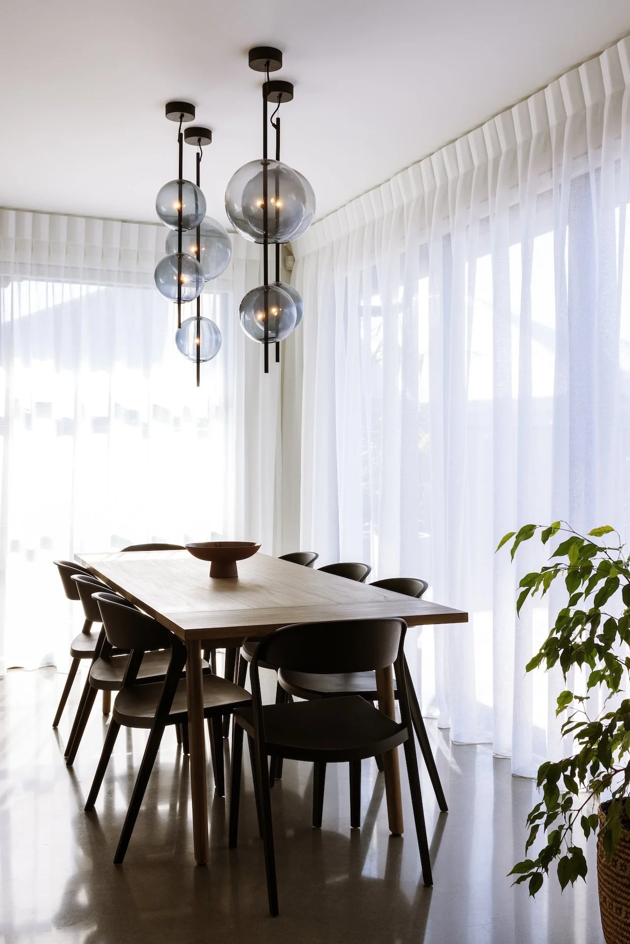 Modern dining room with a rectangular wooden table, six black chairs, a red bowl centerpiece, hanging spherical pendant lights, and large windows with white curtains.