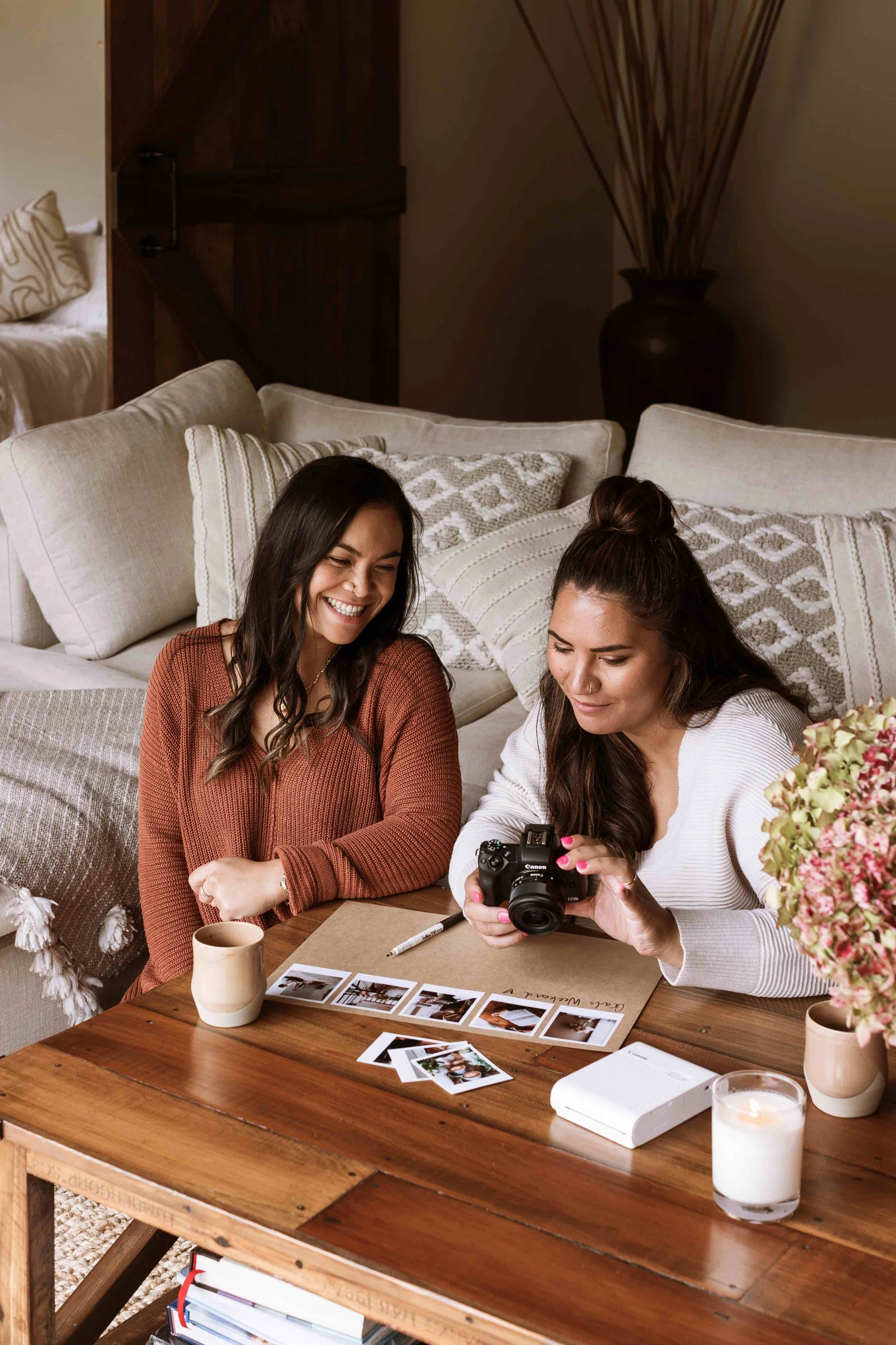 Two women sitting at a wooden coffee table, looking at photographs and a camera with smiles indoors. Lifestyle advertising campaign photography.