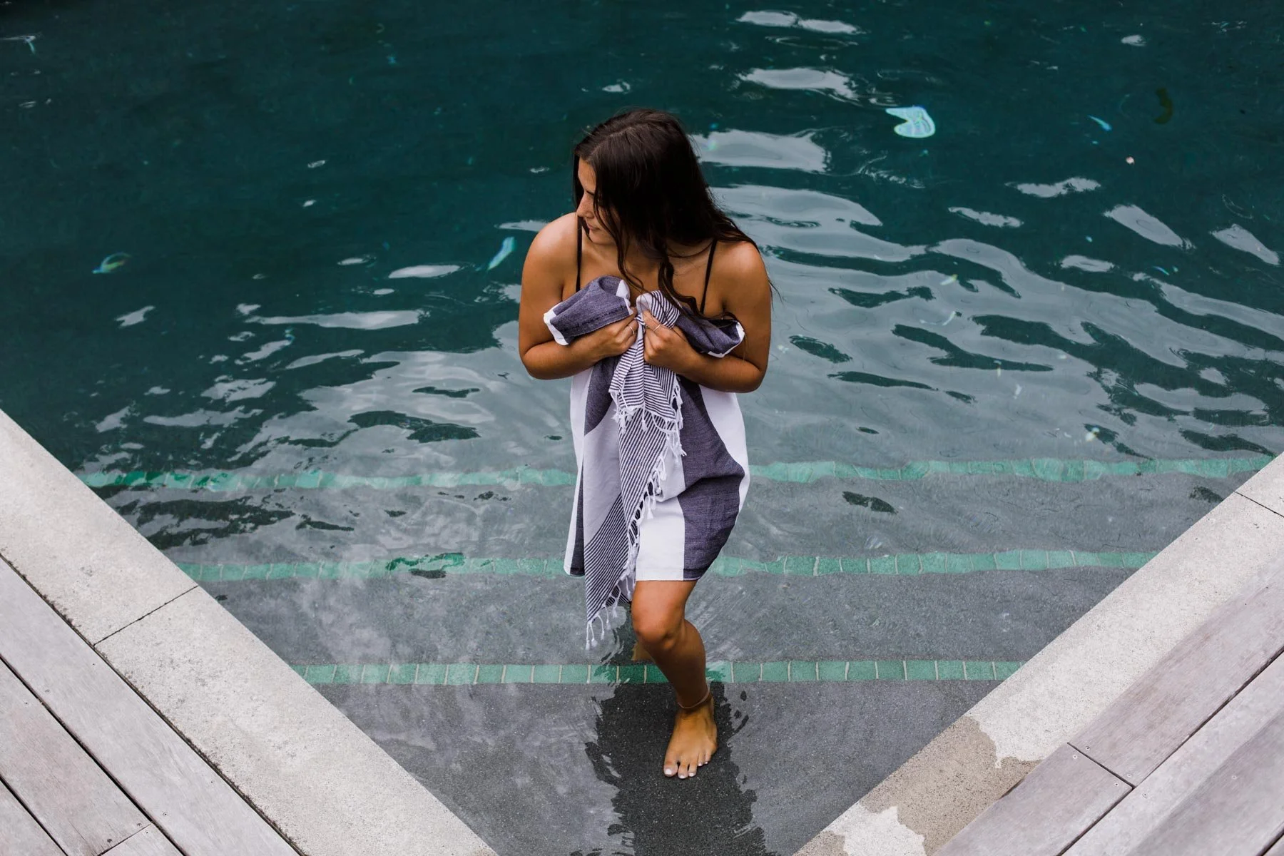 Woman stepping out of a swimming pool, holding a towel against her chest, with wet hair and one foot on the pool step. Photographed for a lifestyle product advertising campaign.