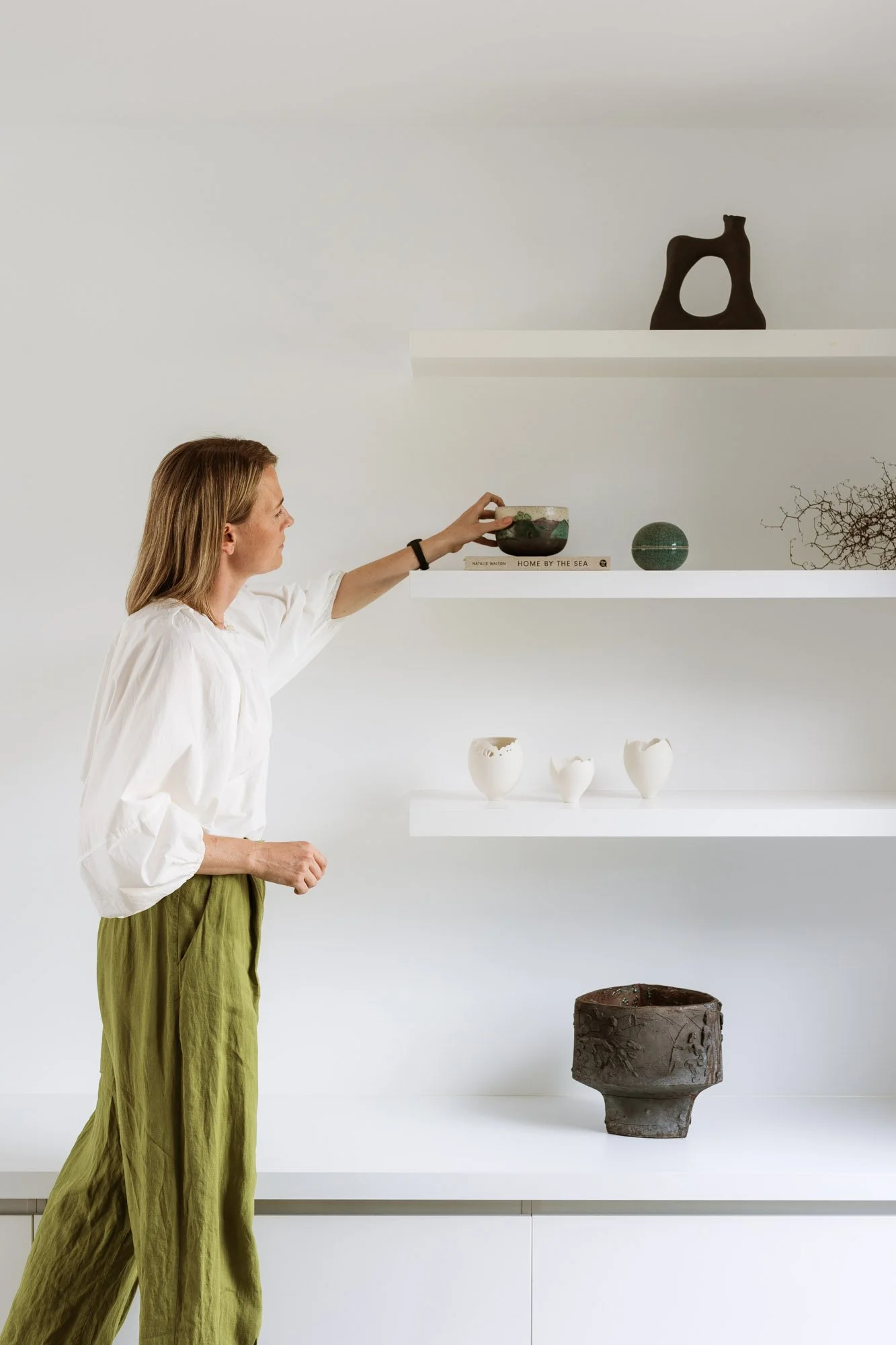 Interior stylist arranging decorative objects on white shelves in a modern, minimalist interior.