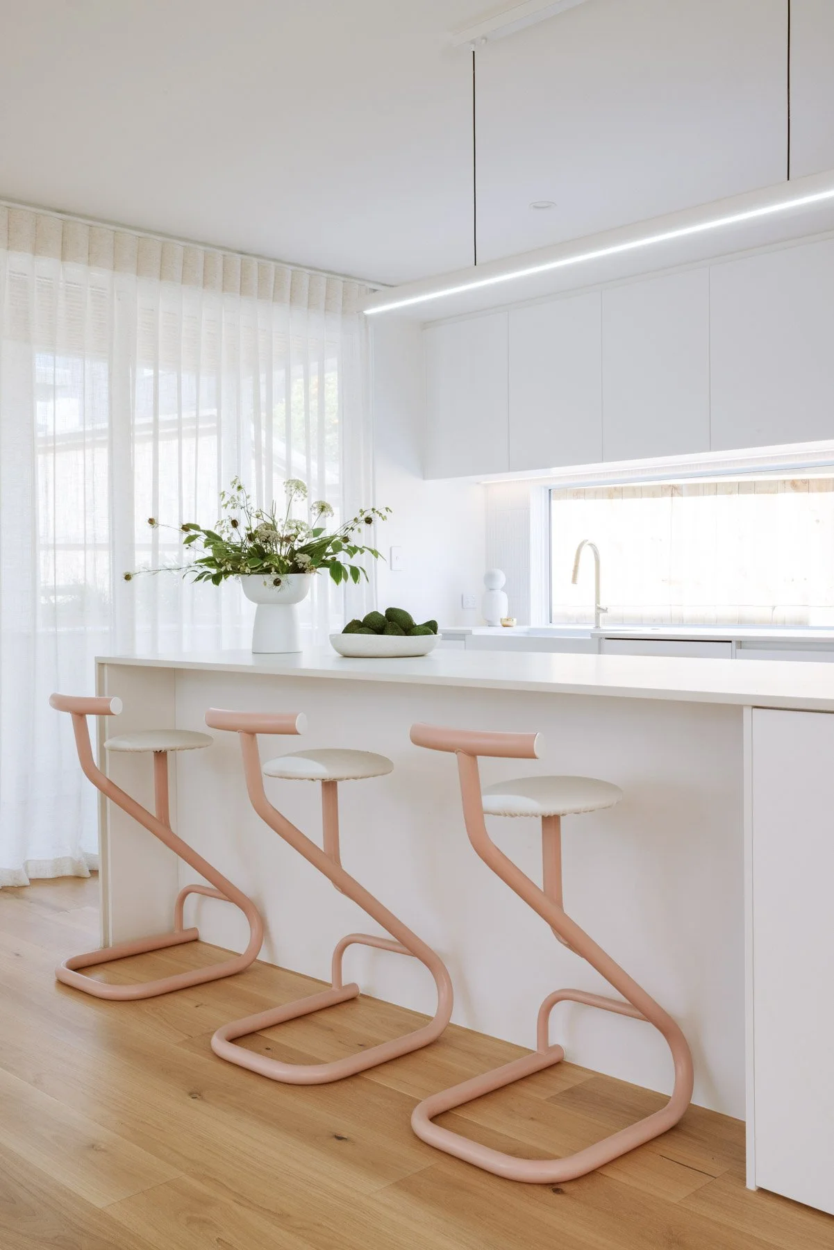 Modern kitchen with white countertops, a window, and three pink bar stools at the counter.
