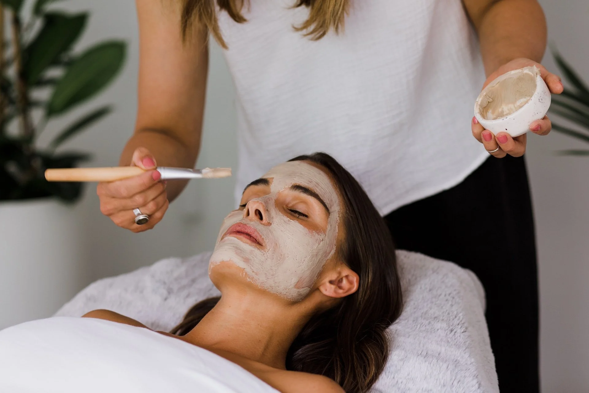 A woman receiving a facial treatment, with a face mask applied, while lying down with eyes closed, and a person applies cream around her forehead with a brush. Lifestyle branding photography for a beauty therapist.