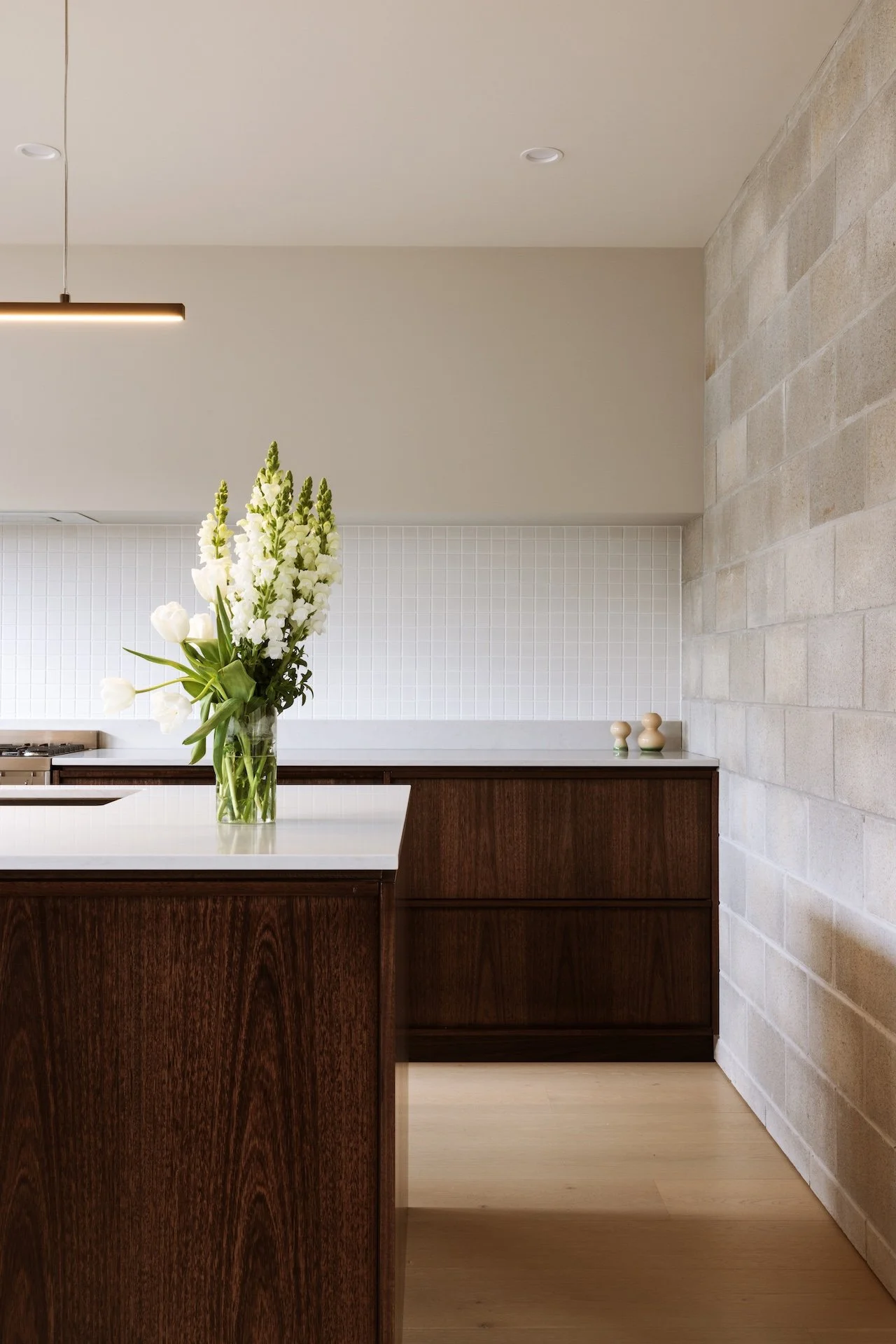 A modern kitchen with a white countertop, dark wooden cabinets, and a large glass vase with white flowers on the counter.