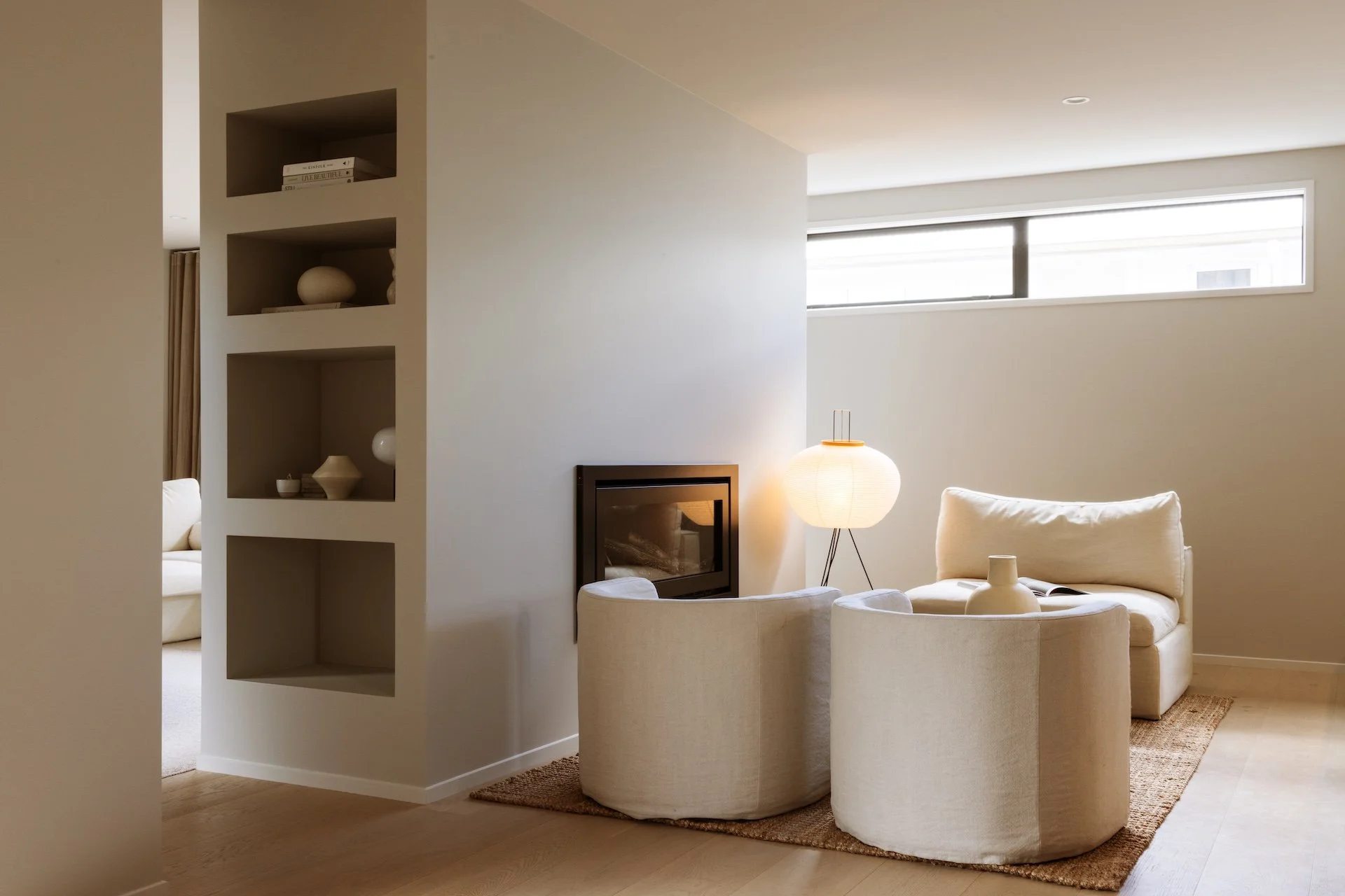 Living room with white sofa, armchair, floor lamp, and a built-in fireplace, featuring minimalist decor and natural light from narrow horizontal window.