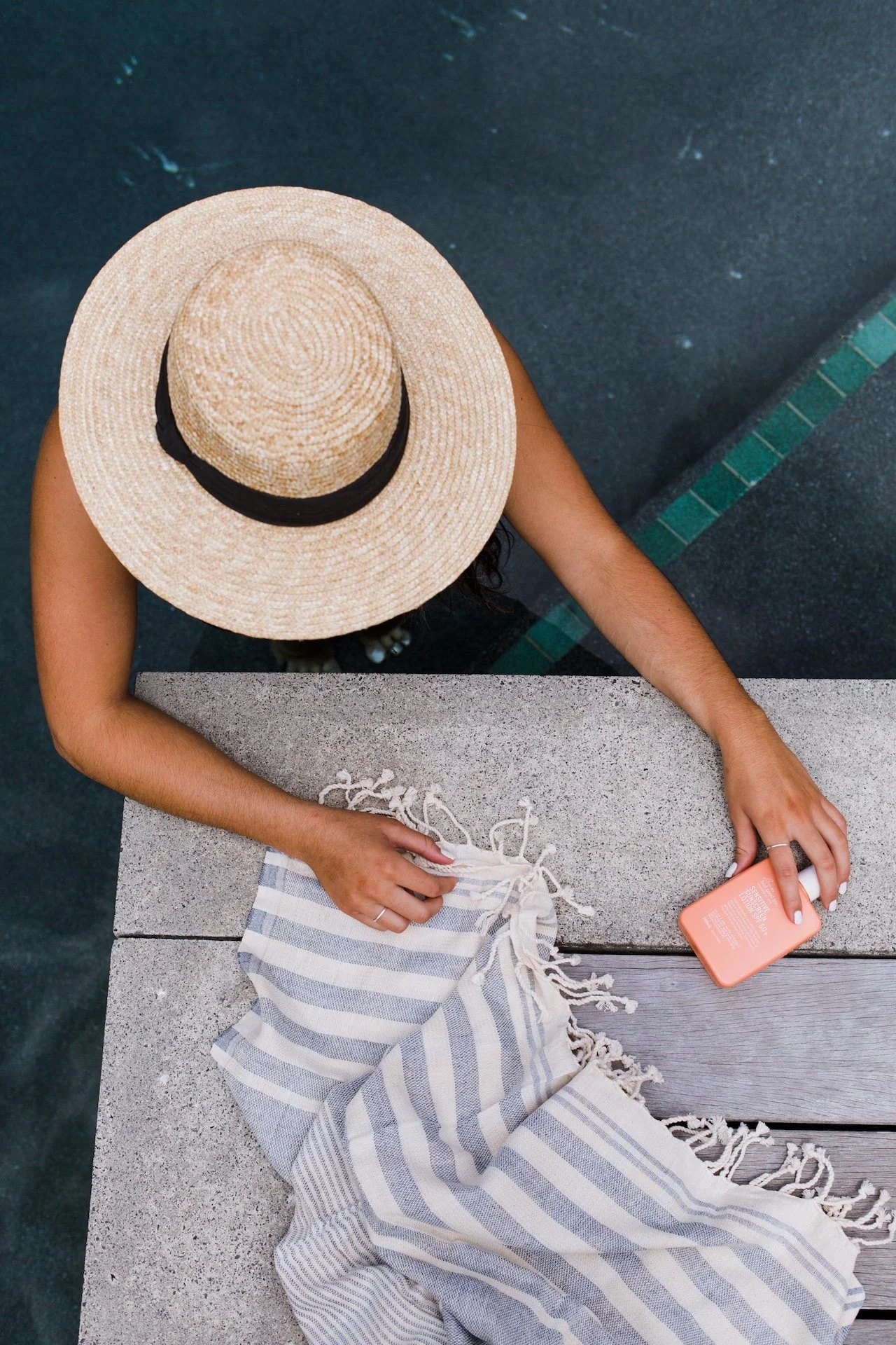 Top-down view of a woman with a large straw hat sitting at a concrete and wooden table, holding a smartphone in one hand and a striped cloth on the table. Lifestyle commercial branding photography in Mount Maunganui.
