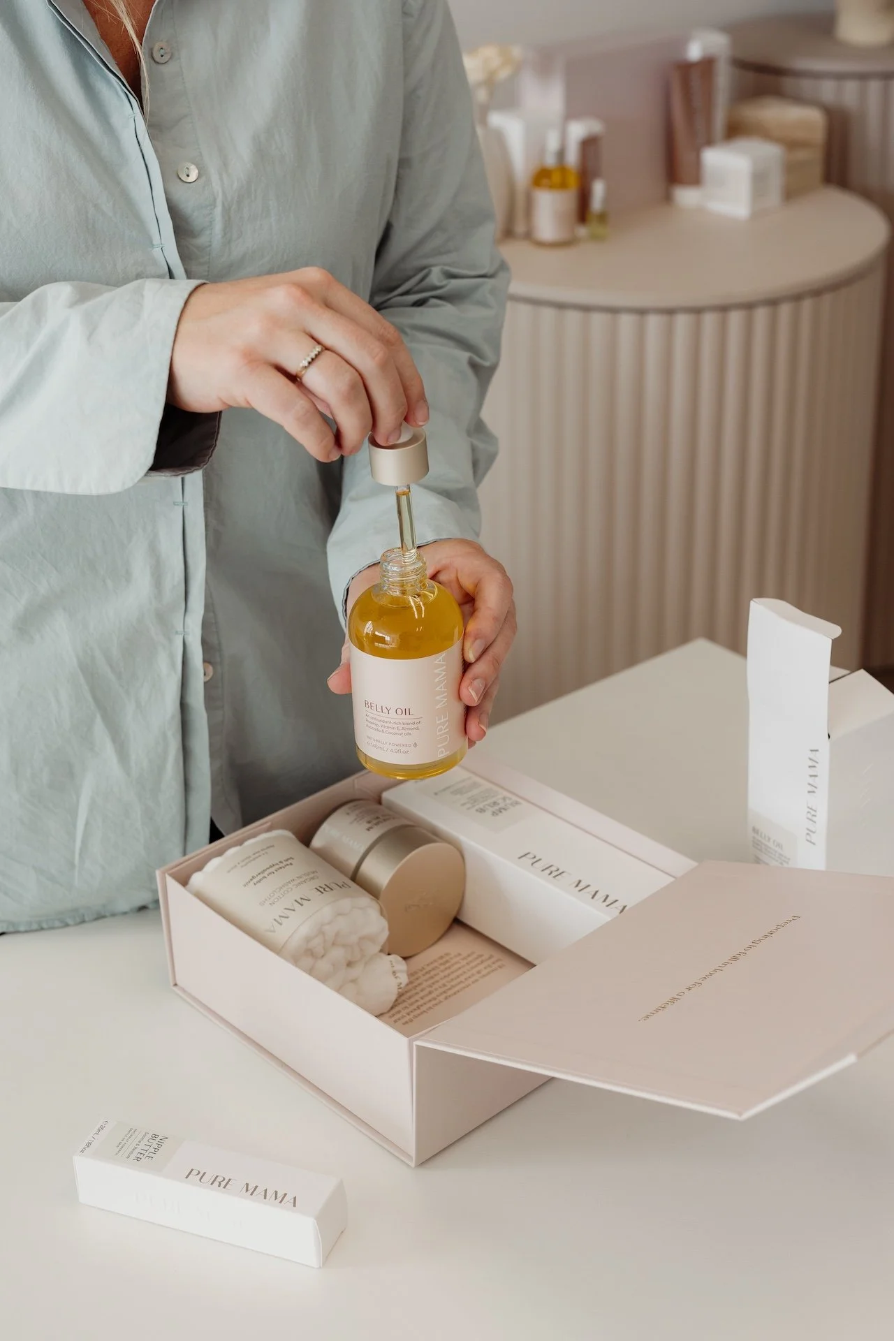 Person holding a bottle of belly oil with a dropper, opening a white box labeled 'Pure Mama' containing skincare products on a white table. Lifestyle commercial branding photography in Mount Maunganui.