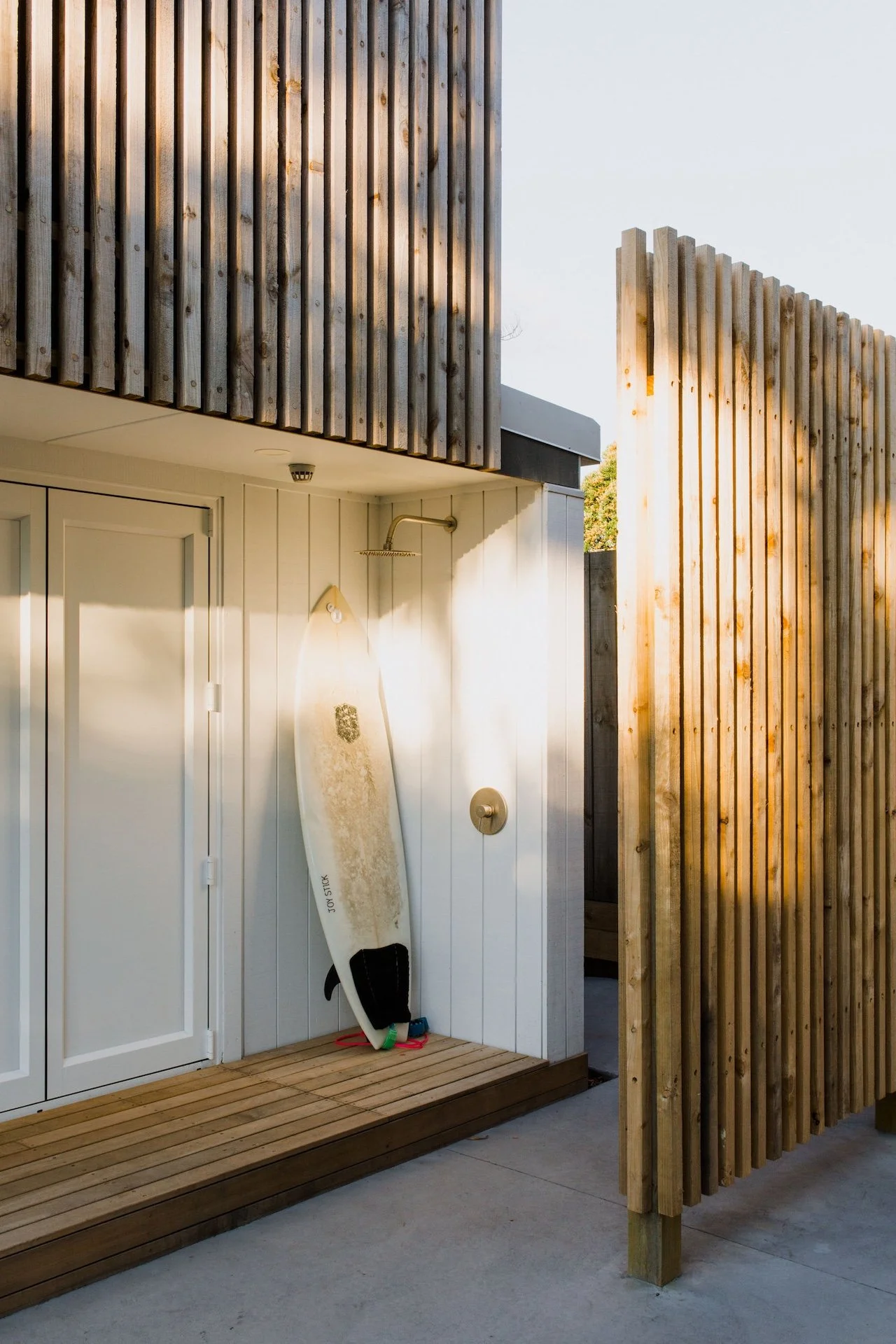Outdoor shower area with a surfboard leaning against the wall, surrounded by wooden fencing.