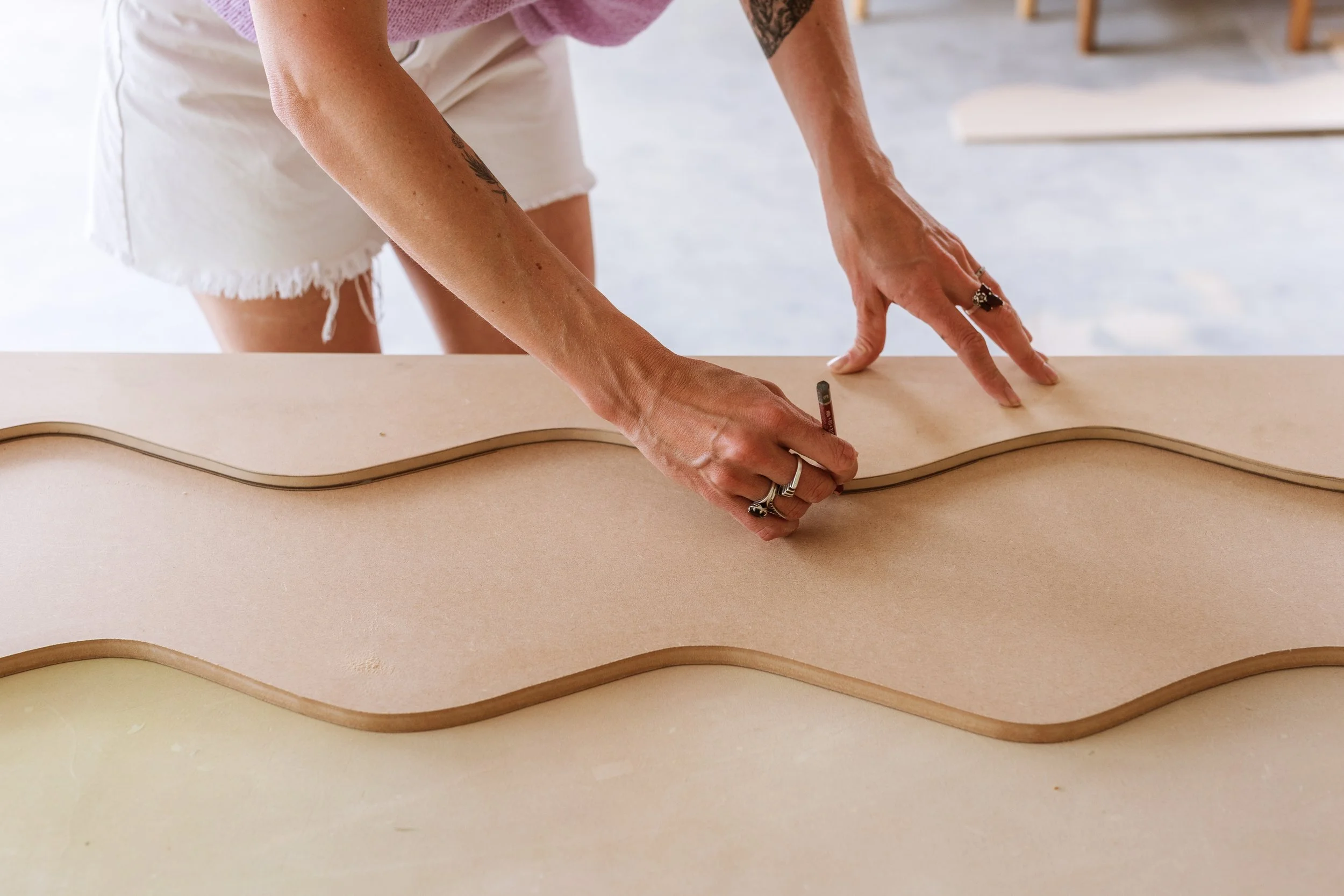 A person is tracing the outline of a wavy-shaped piece of wood on a flat surface, possibly in a woodworking or crafting setting. Lifestyle commercial branding photography in Mount Maunganui.