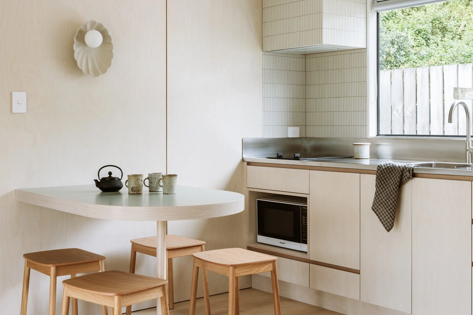 Minimalist kitchen with light wood cabinets, a window above the sink, and a small round dining table with four wooden stools. A teapot and cups are on the table, and a towel hangs over the sink.