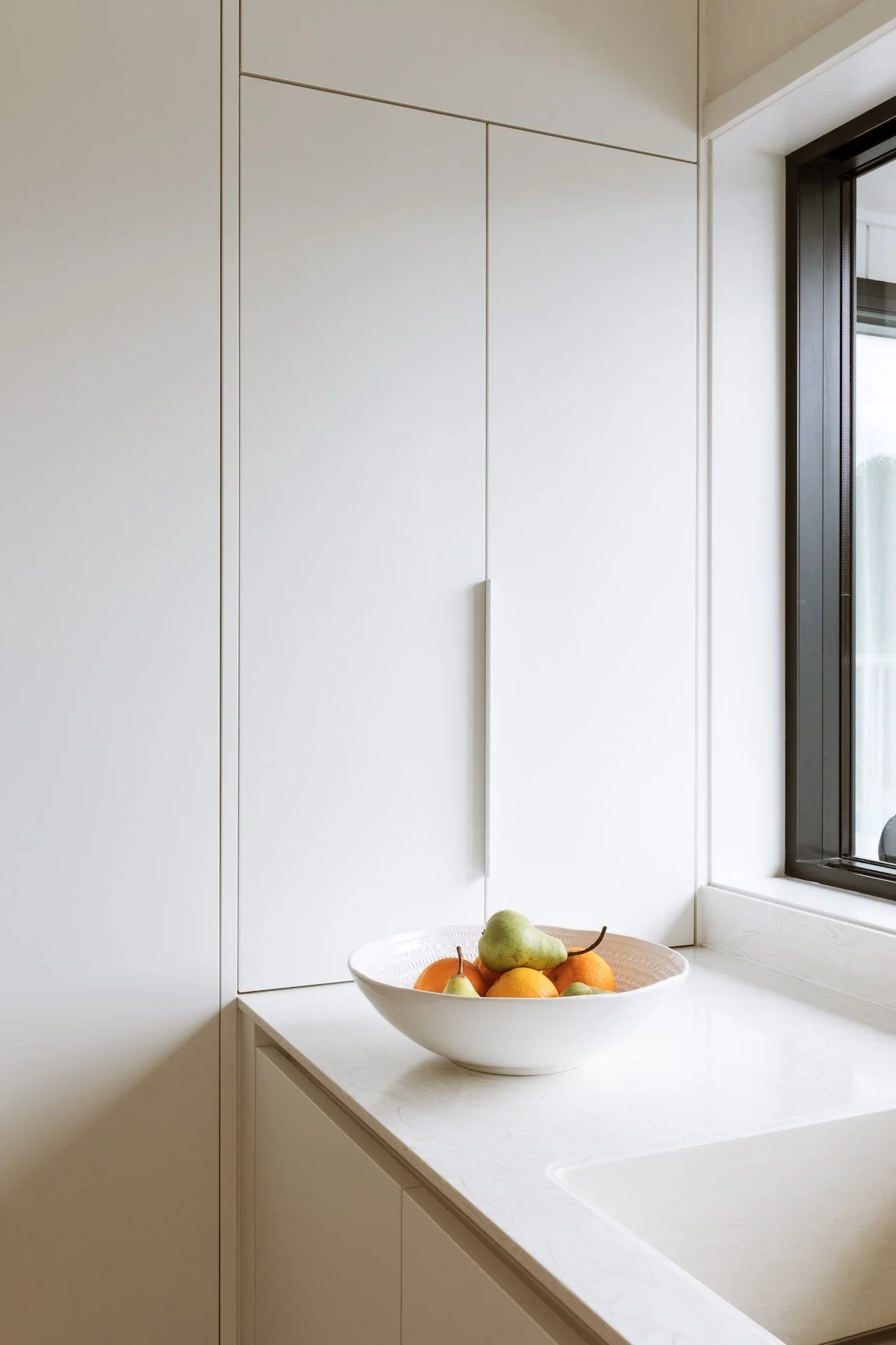 A white bowl filled with pears, oranges, and other fruits sits on a white kitchen countertop near a window with black framing.