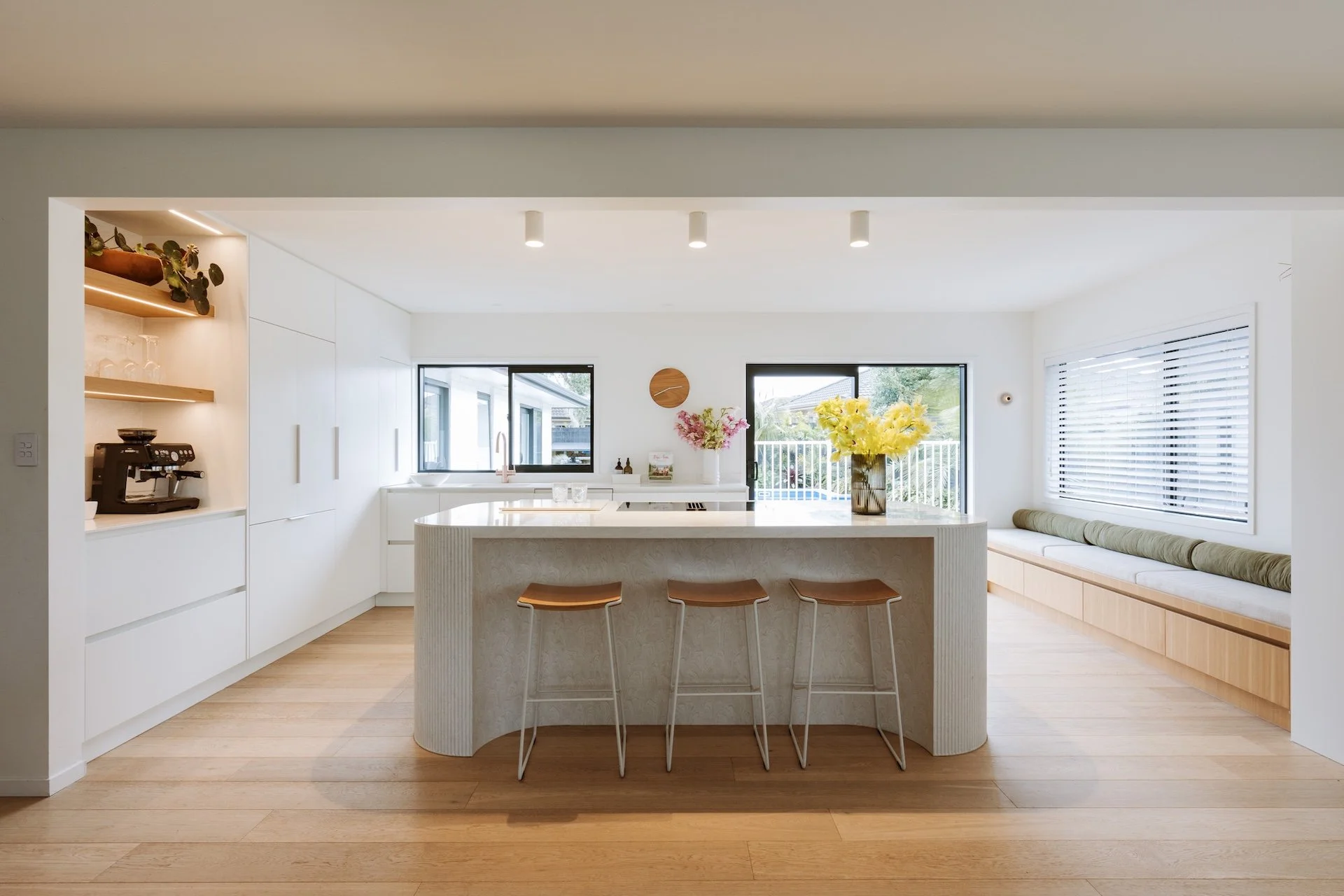 Modern kitchen with white cabinets, a curved island with three wooden stools, large windows, a built-in bench with green cushions, and fresh flowers on the counter.