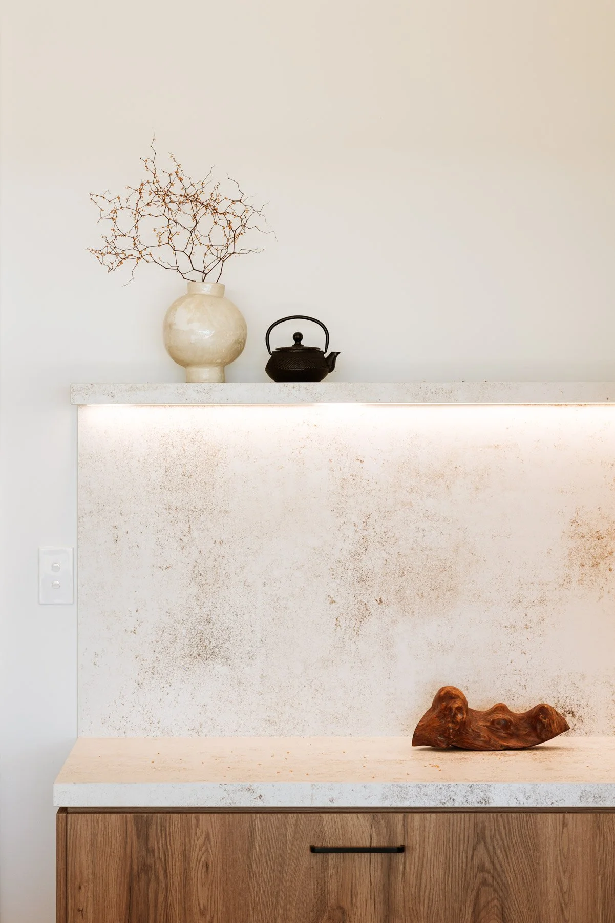 Minimalist kitchen counter with a dried tree branch in a beige ceramic vase, a black teapot, and a wooden decorative piece on the counter, with white walls and wooden cabinets.
