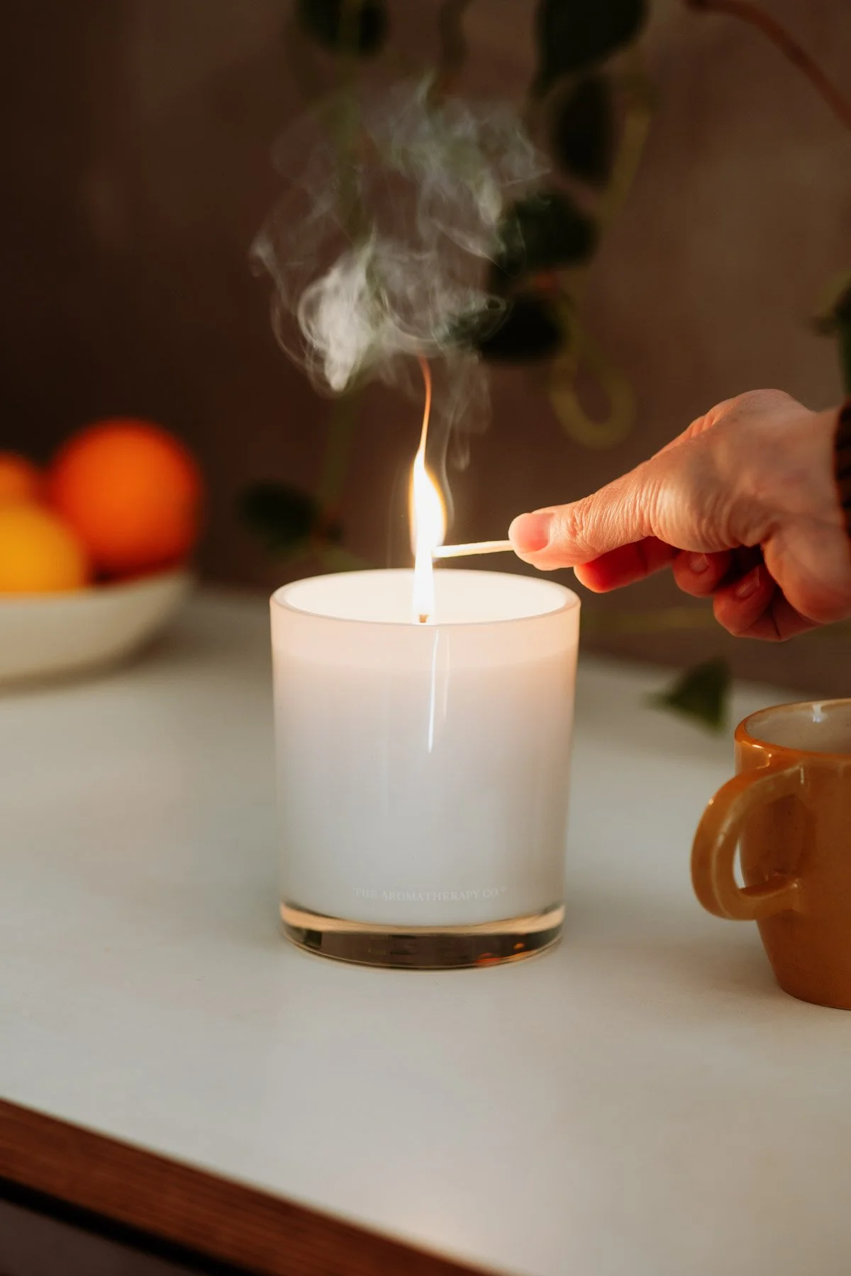 A person stirs a lit white candle with a matchstick, with smoke rising from the flame, on a white surface with a mug nearby and blurred oranges in the background. Professional Lifestyle Product photography.
