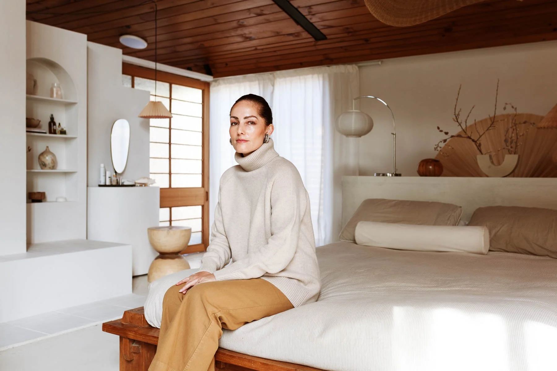 A woman sitting on the edge of a bed in a cozy, minimalist bedroom with natural light and wooden accents for Assembly Label campaign.