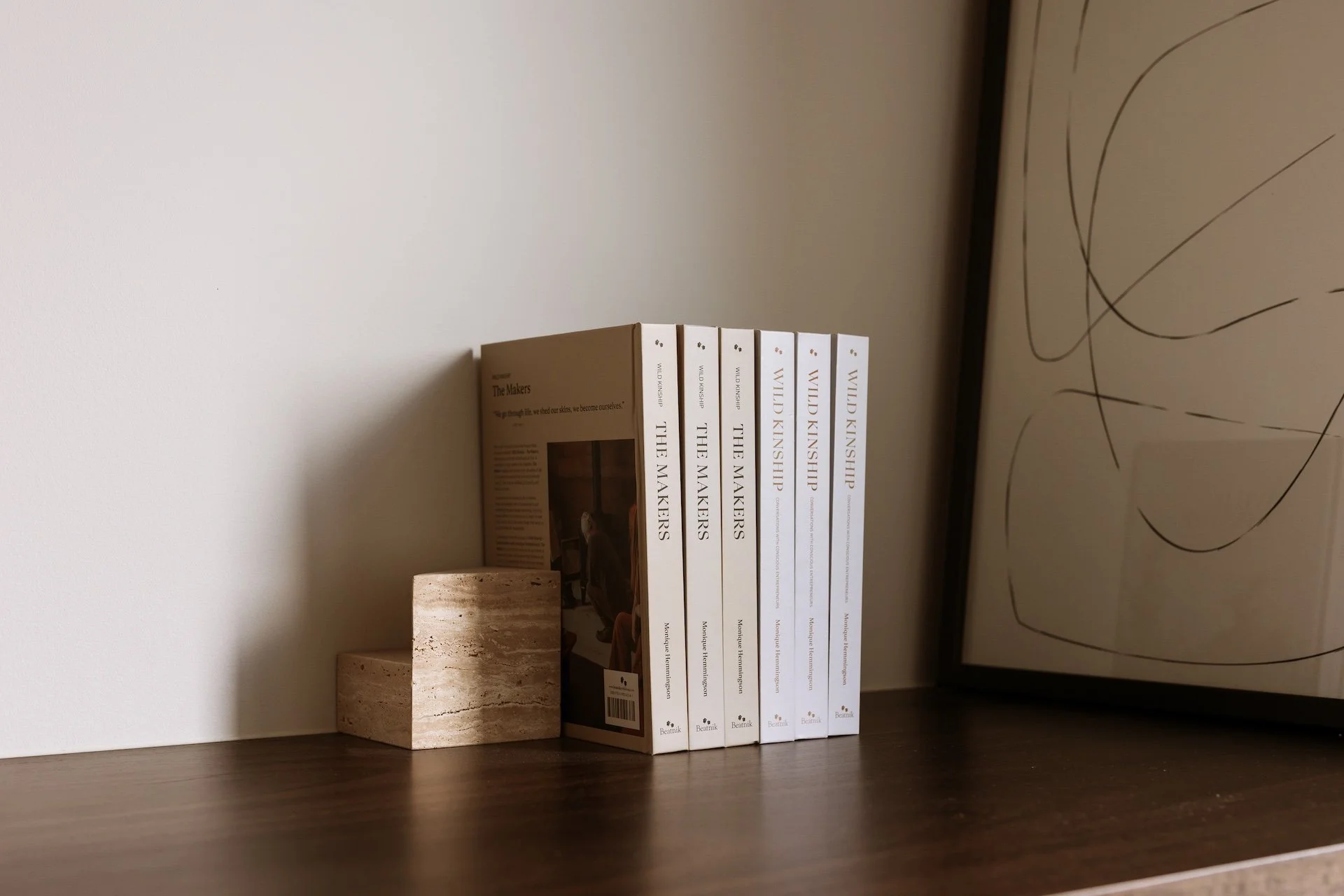 A set of four books titled 'The Makers' by Wild Kinship placed on a wooden surface, next to a small, curved marble block, with a framed abstract drawing partially visible on the right side.