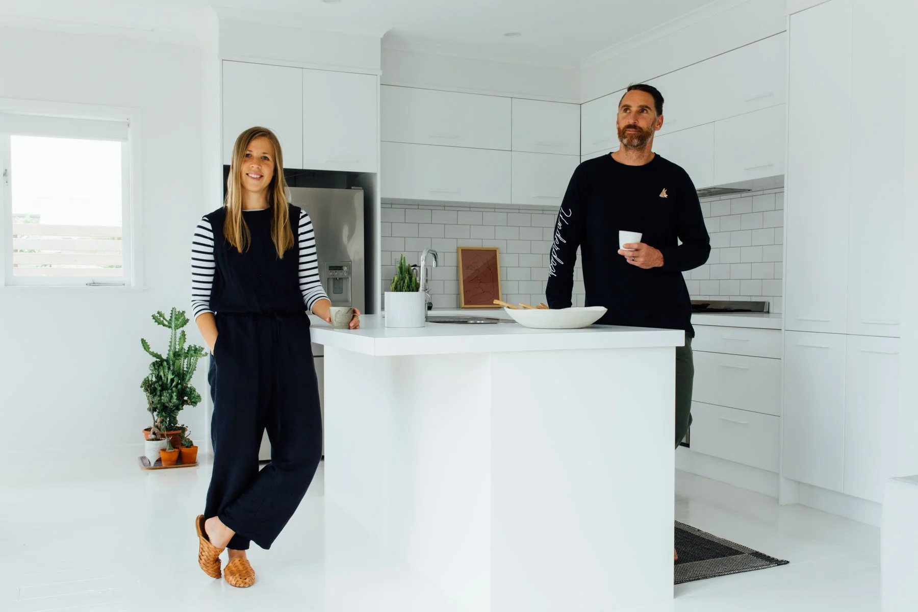 A woman and a man are standing in a bright, white kitchen. The woman is smiling, holding a mug, and leaning against a kitchen island. The man is holding a white mug. Photographed to showcase the architectural and design elements of the owners' home.