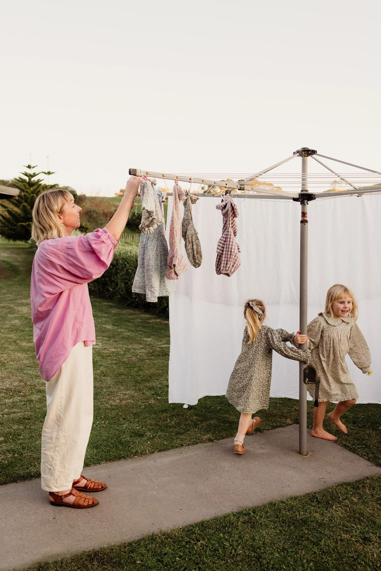 A woman hanging laundry on a rotary clothesline while two young girls play nearby outdoors, with a background of trees and a grassy field.