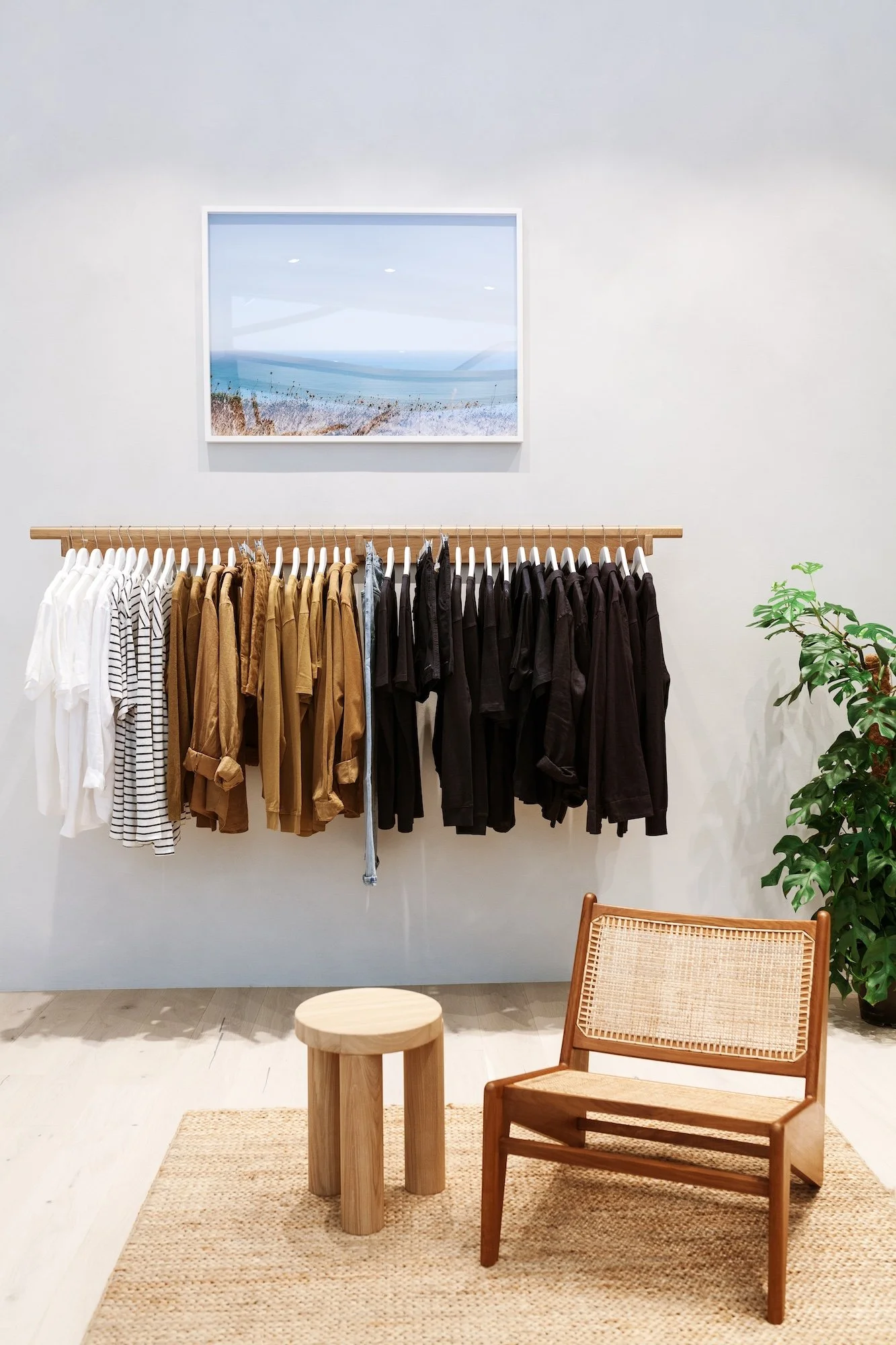 Clothing display in a boutique, with white, striped, brown, black shirts hanging on a wooden rack. Beneath the rack is a beige woven rug, a round wooden stool, and a wooden chair with a woven backrest. To the right is a potted plant with green foliag
