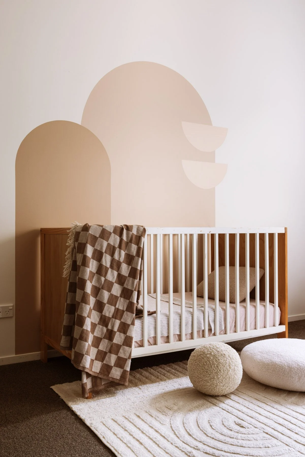 A nursery with a white crib, beige and brown checkered blanket, textured pillow, and a decorative ball on a cream rug, with a beige and pink abstract wall mural.