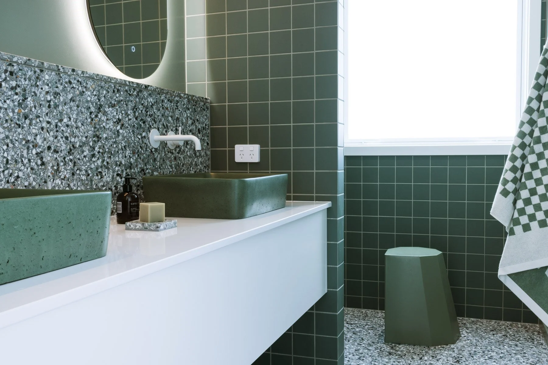 Modern bathroom with green square tiled walls, terrazzo countertop, green vessel sinks, round mirror, white wall-mounted faucet, black soap dispenser, candle, green geometric stool, and checkered towel near a window.