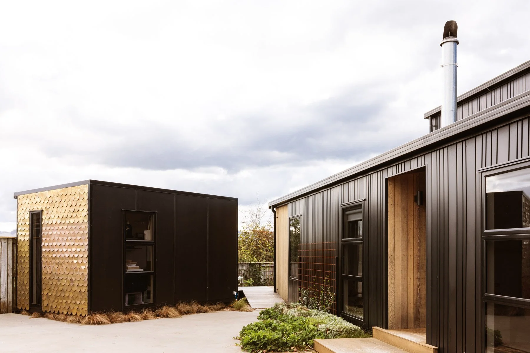 Modern exterior of an architecturally designed home and studio with black and gold siding and black framed windows, small landscaped garden in front, cloudy sky overhead.