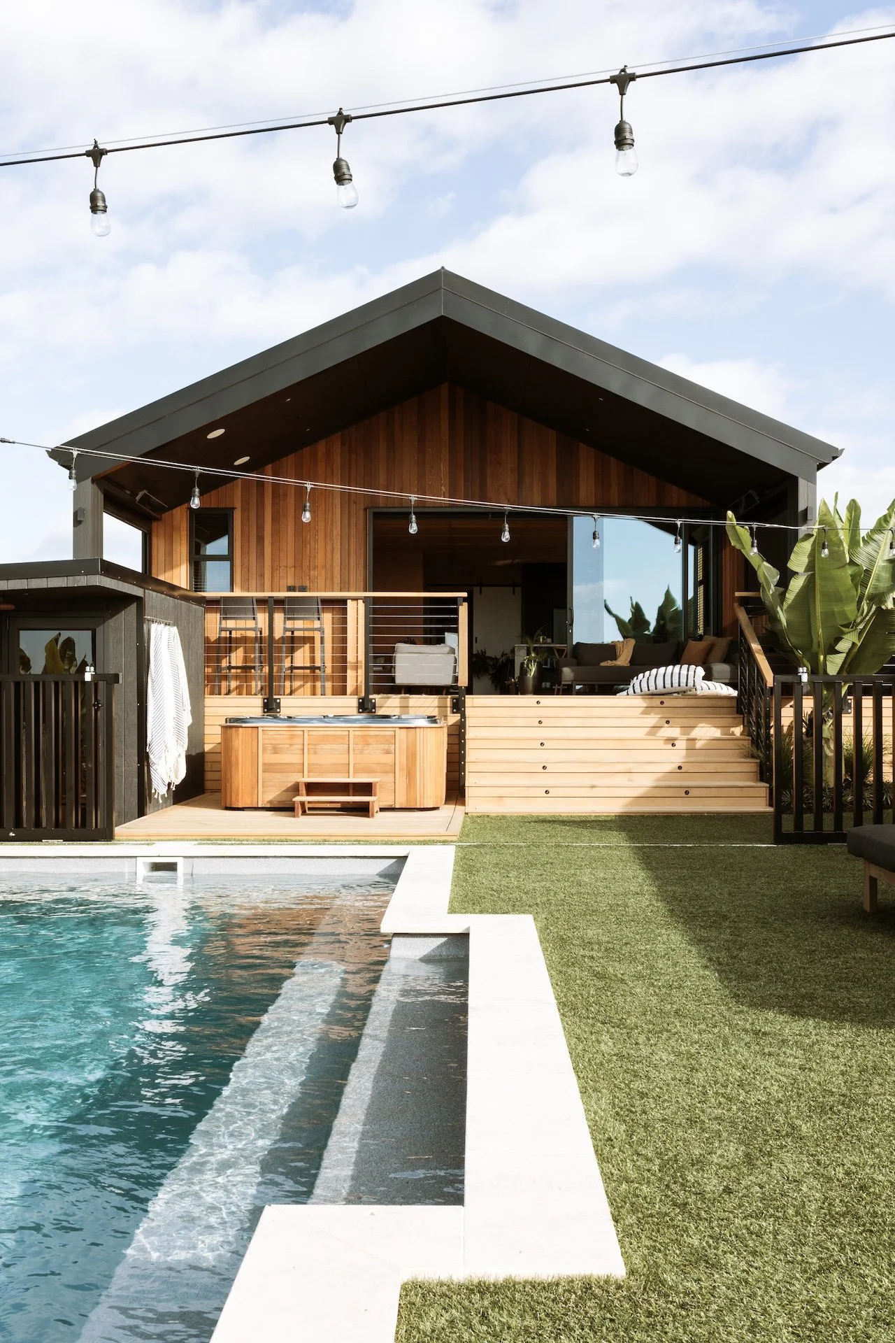 Modern backyard with a swimming pool, wooden deck, hot tub, and a two-story architecturally designed house with a dark roof and large windows, surrounded by string lights and tropical plants.