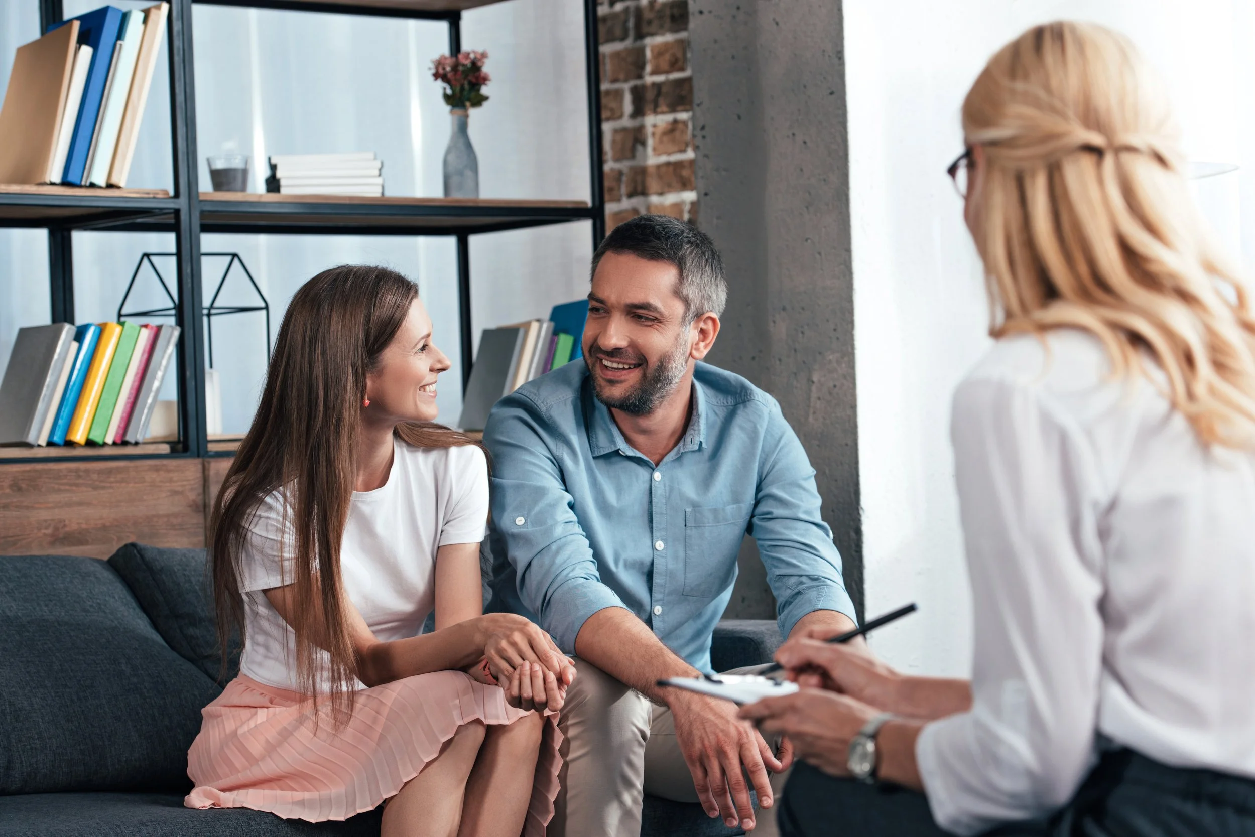 A couple sitting on a sofa at a therapy session, holding hands and smiling, facing a therapist with blonde hair and glasses holding a clipboard, in a modern room with a bookshelf and brick wall.