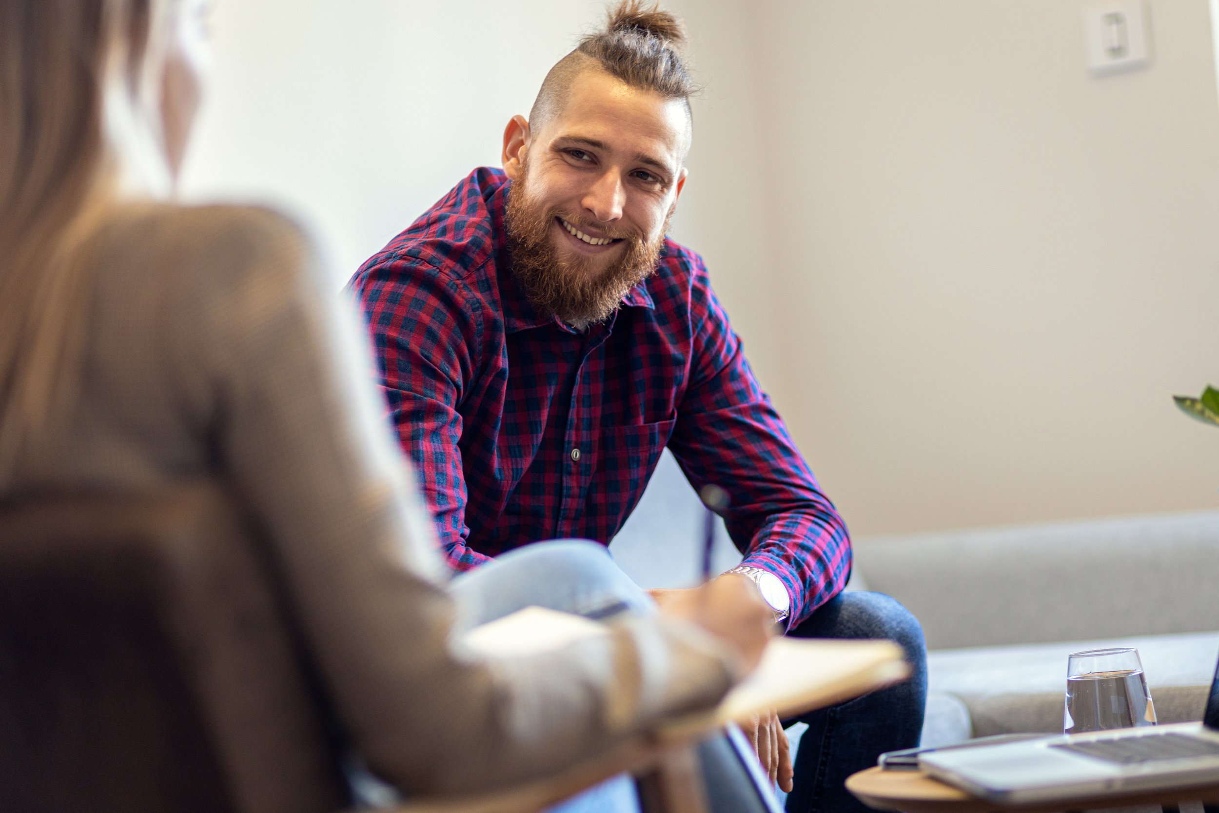 A man with a beard and long hair tied up in a bun, wearing a red and blue plaid shirt, smiling and sitting at a table during a meeting or discussion.