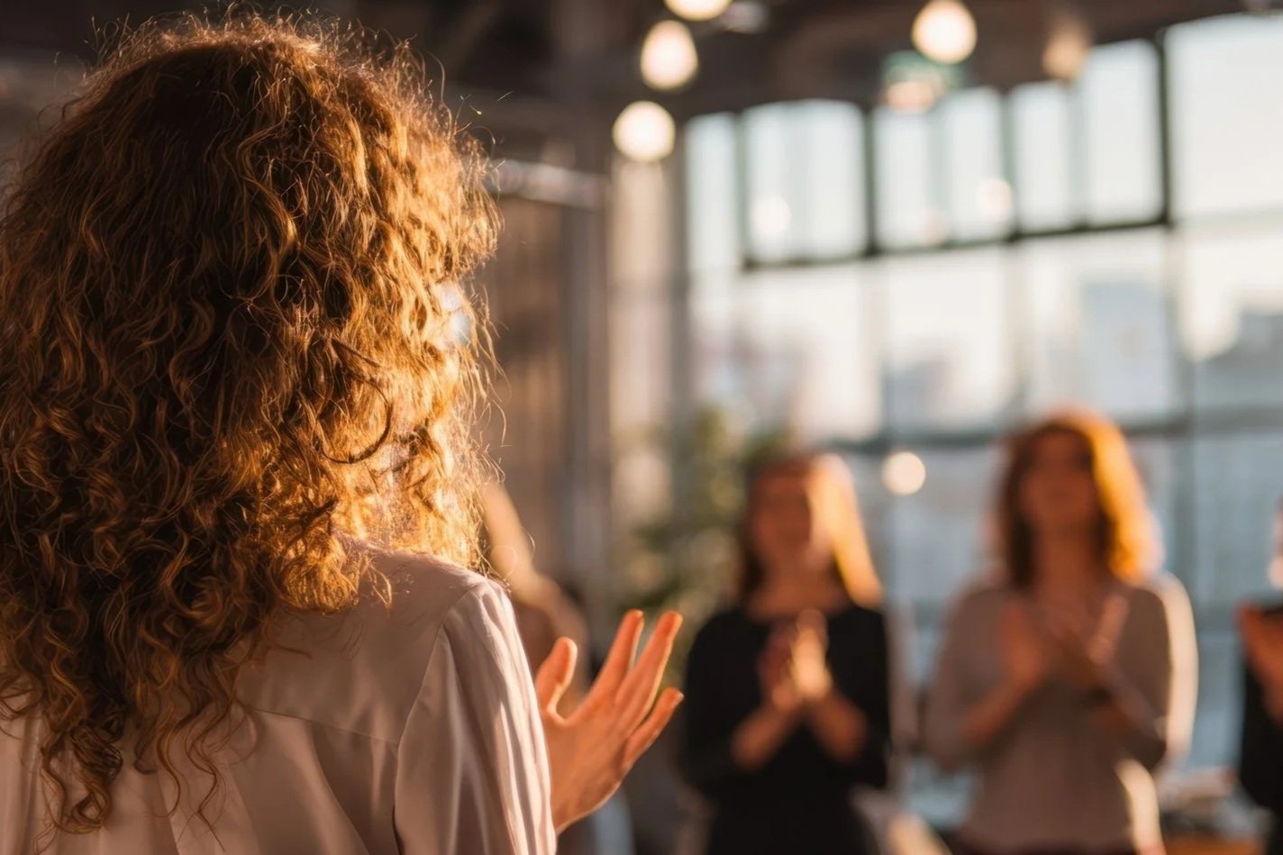 Group of women clapping in a warmly lit indoor setting with large windows.