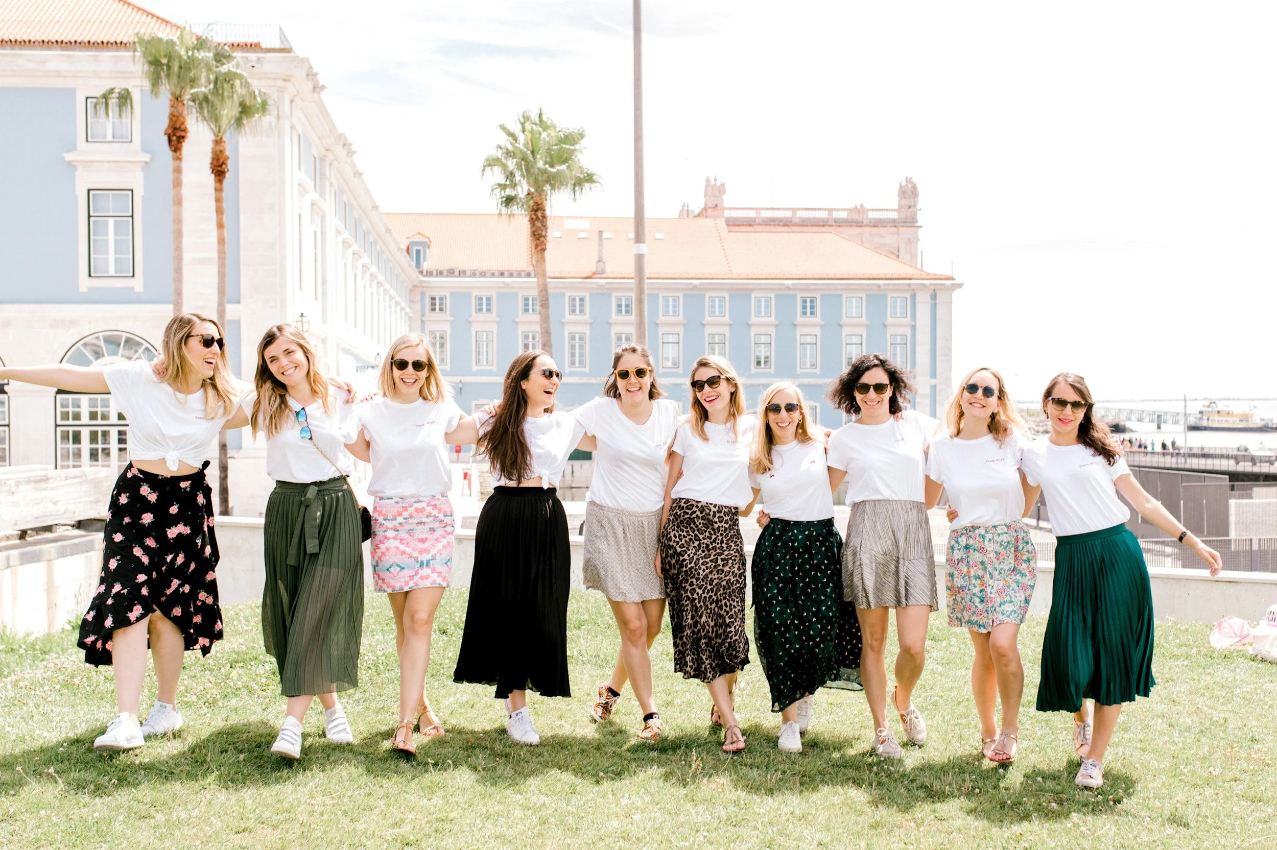 A group of eleven women walking arm-in-arm on a grassy area outdoors, smiling and wearing casual white t-shirts and colorful skirts, with a cityscape and palm trees in the background supporting HER Society.