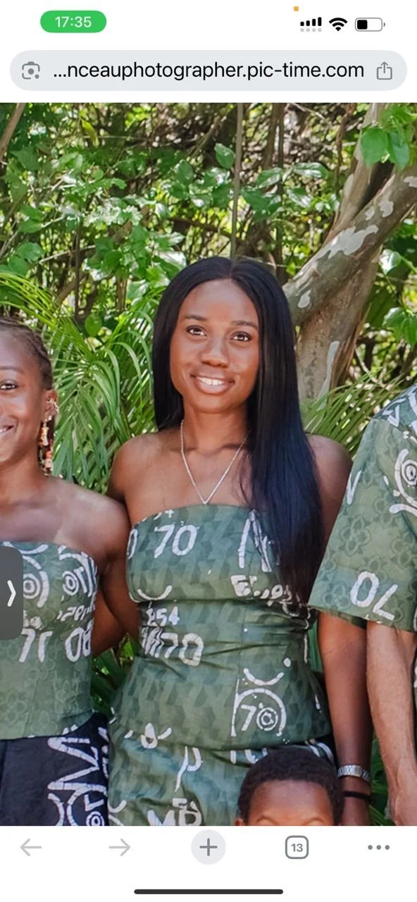 A woman smiling, wearing a green strapless dress with white patterns, standing outdoors with green foliage and trees in the background.