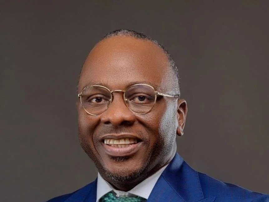A man wearing glasses a green tea and blue blazer against a dark background