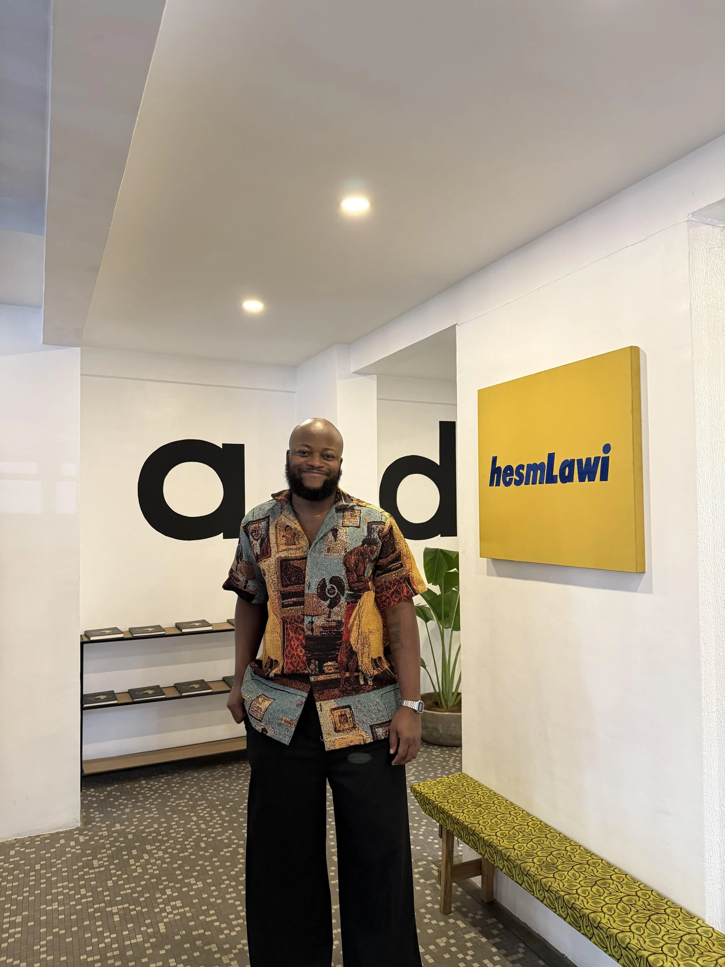 A man posing in a hall way, standing up. Wearing a colourful button up shirt and dark trousers