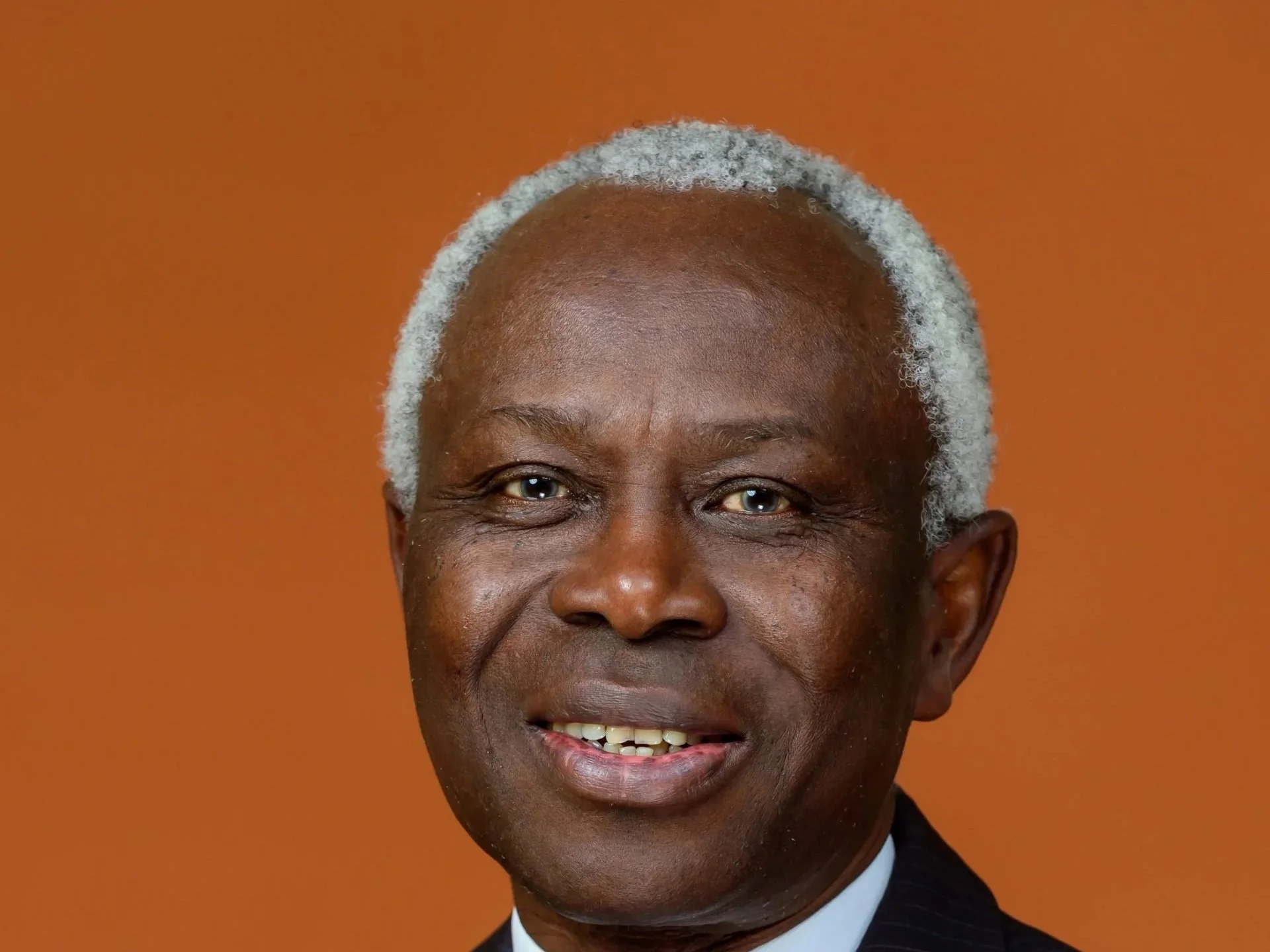 A man with grey hair wearing a blazer and bright blue tie, posing against a burnt orange background.