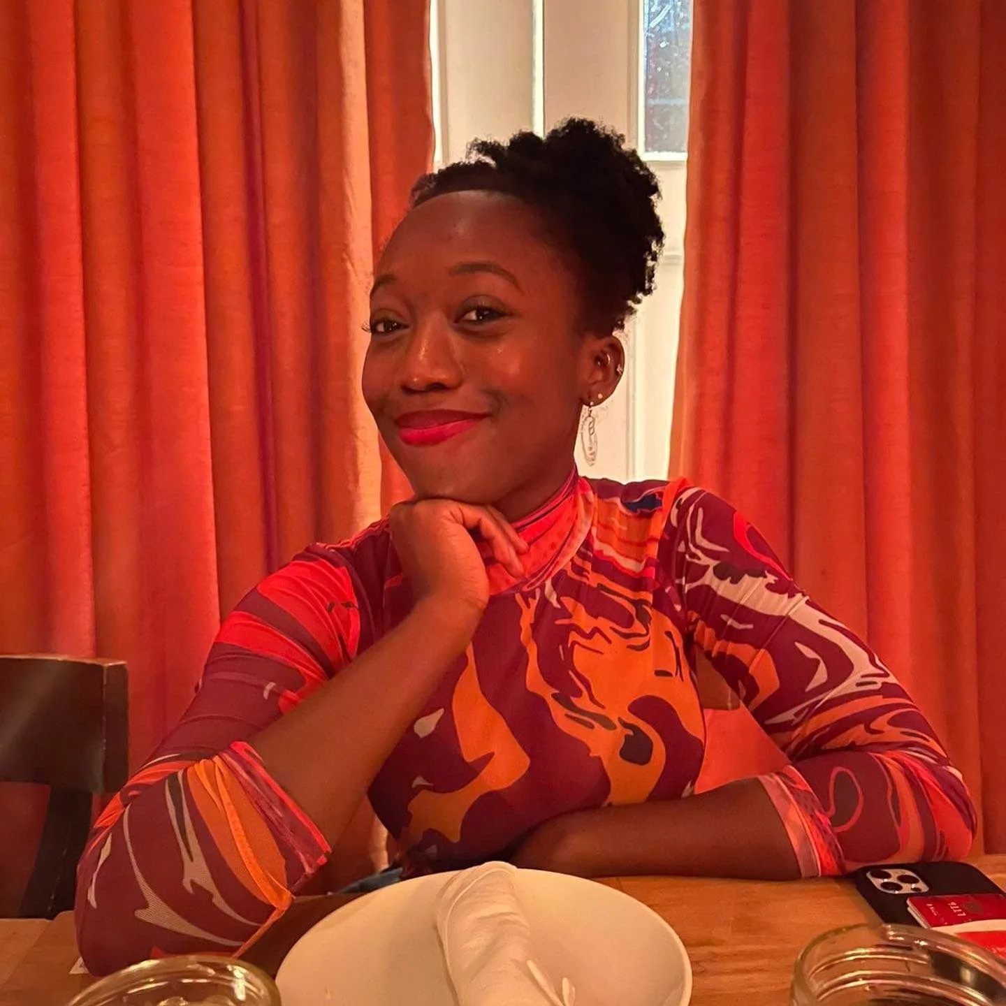 A woman with dark skin and curly hair styled in an updo, smiling and resting her chin on her hand at a table with a white plate and glasses, in a room with orange curtains and a window.