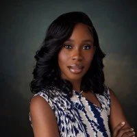 A woman with dark, wavy hair wearing a patterned sleeveless top, posing against a dark background.