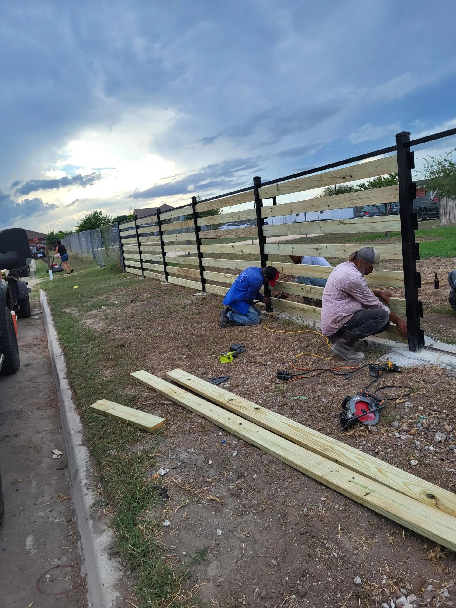 People building a wooden fence on a sidewalk in an outdoor area during the daytime with a cloudy sky.