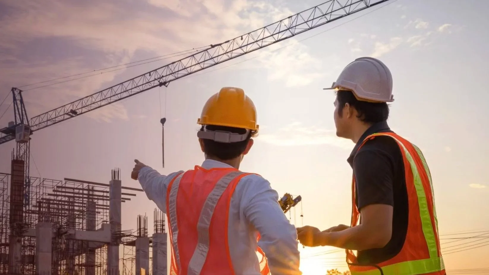 Two construction workers wearing safety vests and helmets standing at a construction site with a crane in the background during sunset, one pointing towards the site.