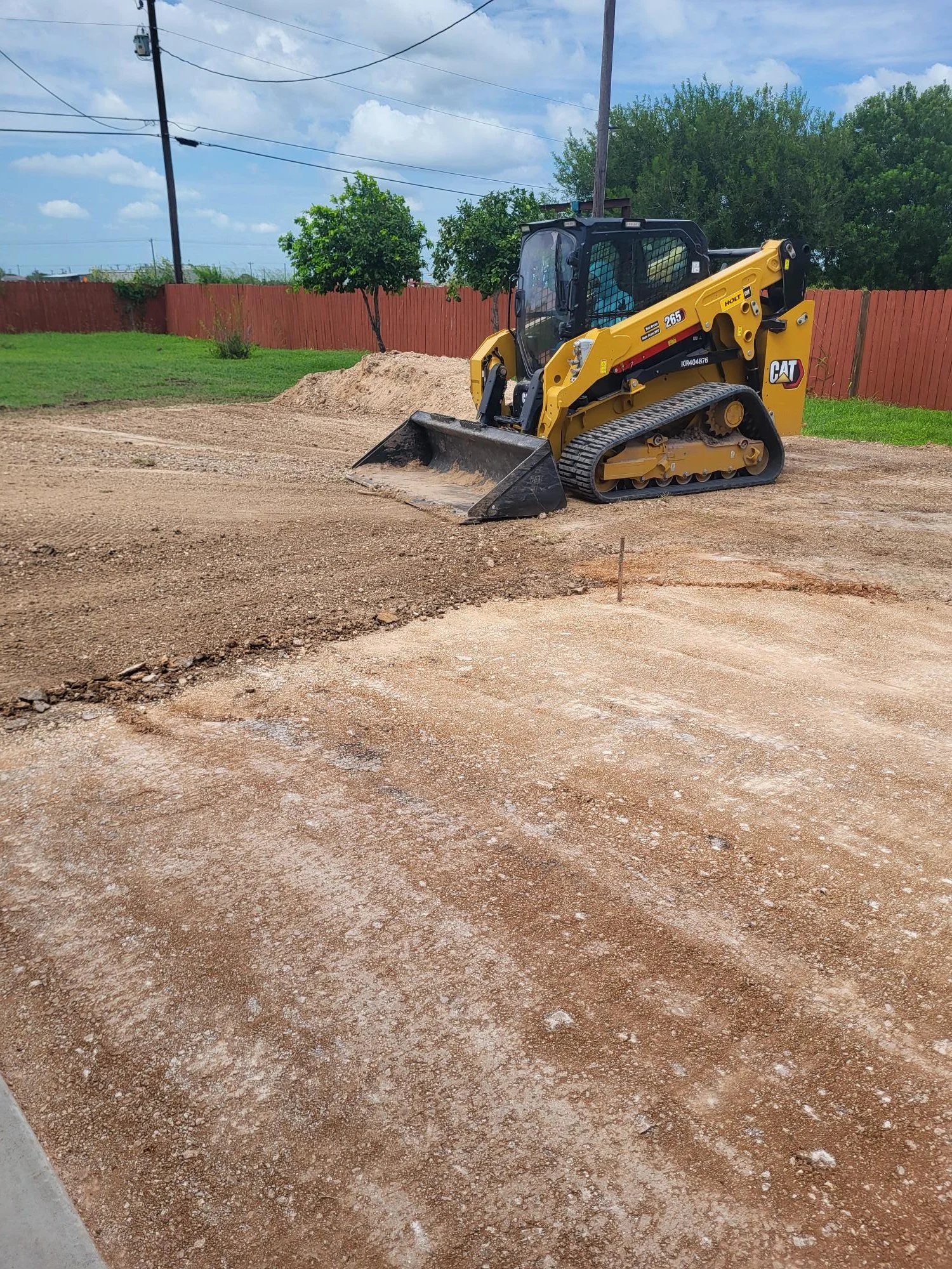 A yellow Caterpillar compact tracked loader with a black bucket attachment working on a dirt surface outdoors, fencing and trees in the background under a partly cloudy sky.
