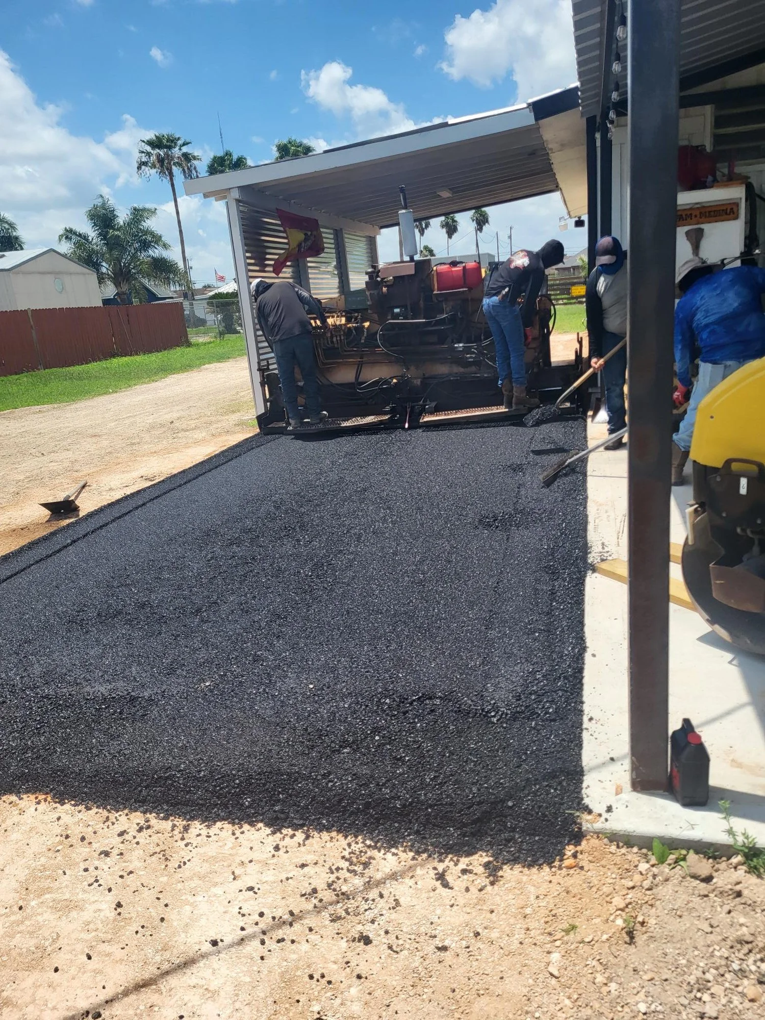 Workers laying fresh asphalt in front of a building on a sunny day with palm trees in the background.