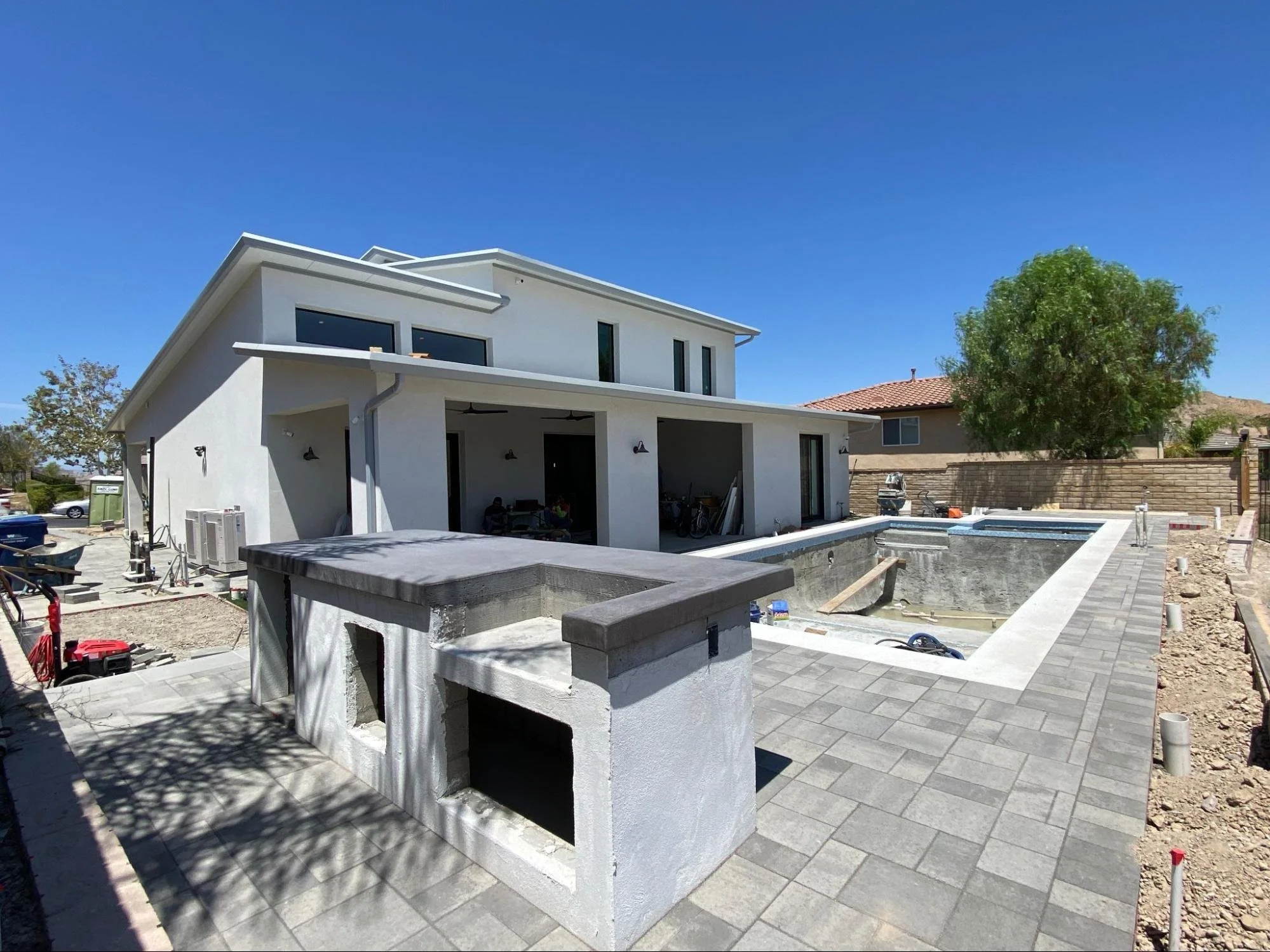 Under construction modern two-story house with a backyard pool area, surrounded by a patio with gray pavers, on a bright sunny day with clear blue skies and a large tree in the background.