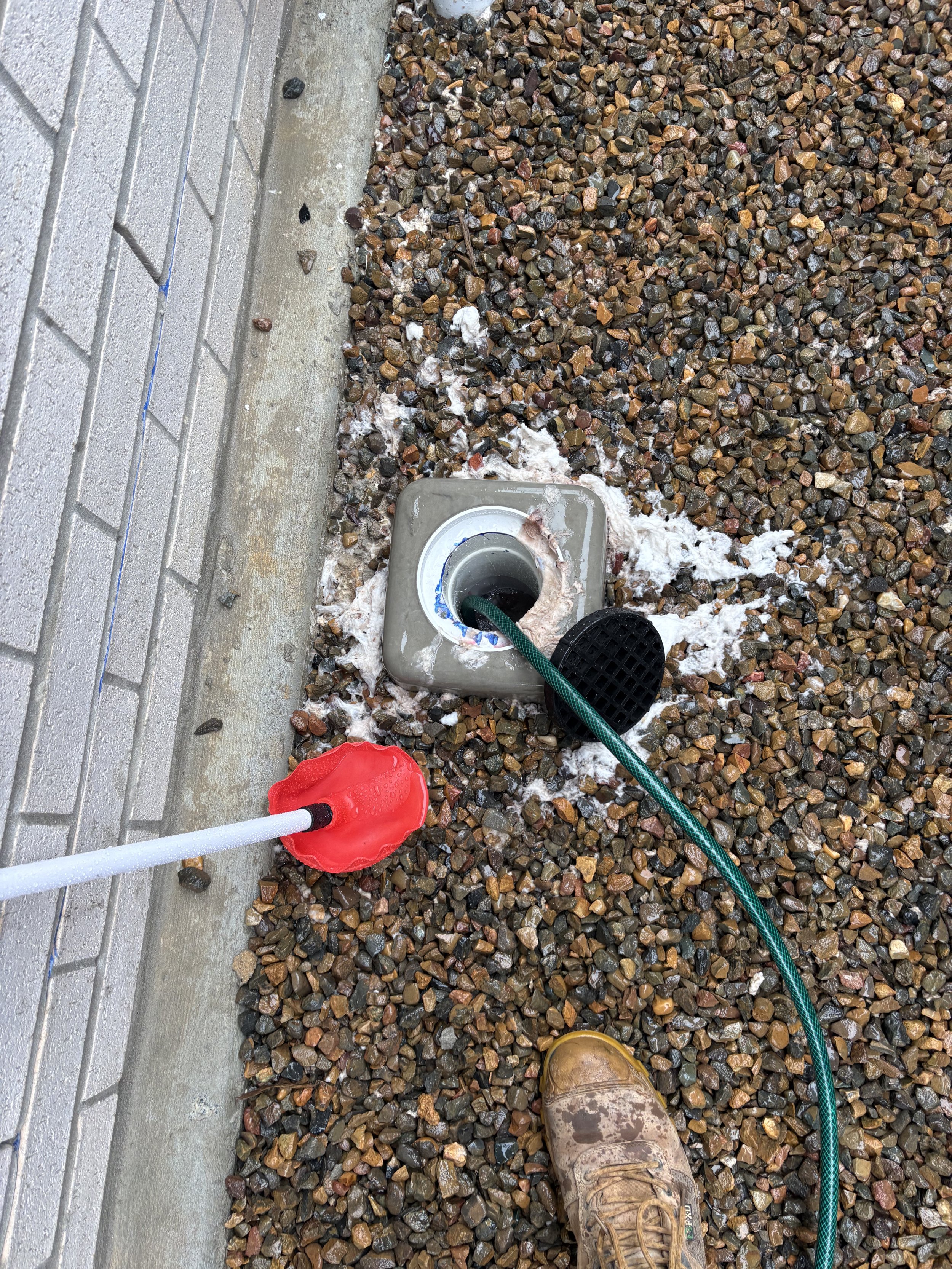 Person wearing muddy work boots installing a water drain or pipe in a gravel and brick patio area during construction or maintenance work.