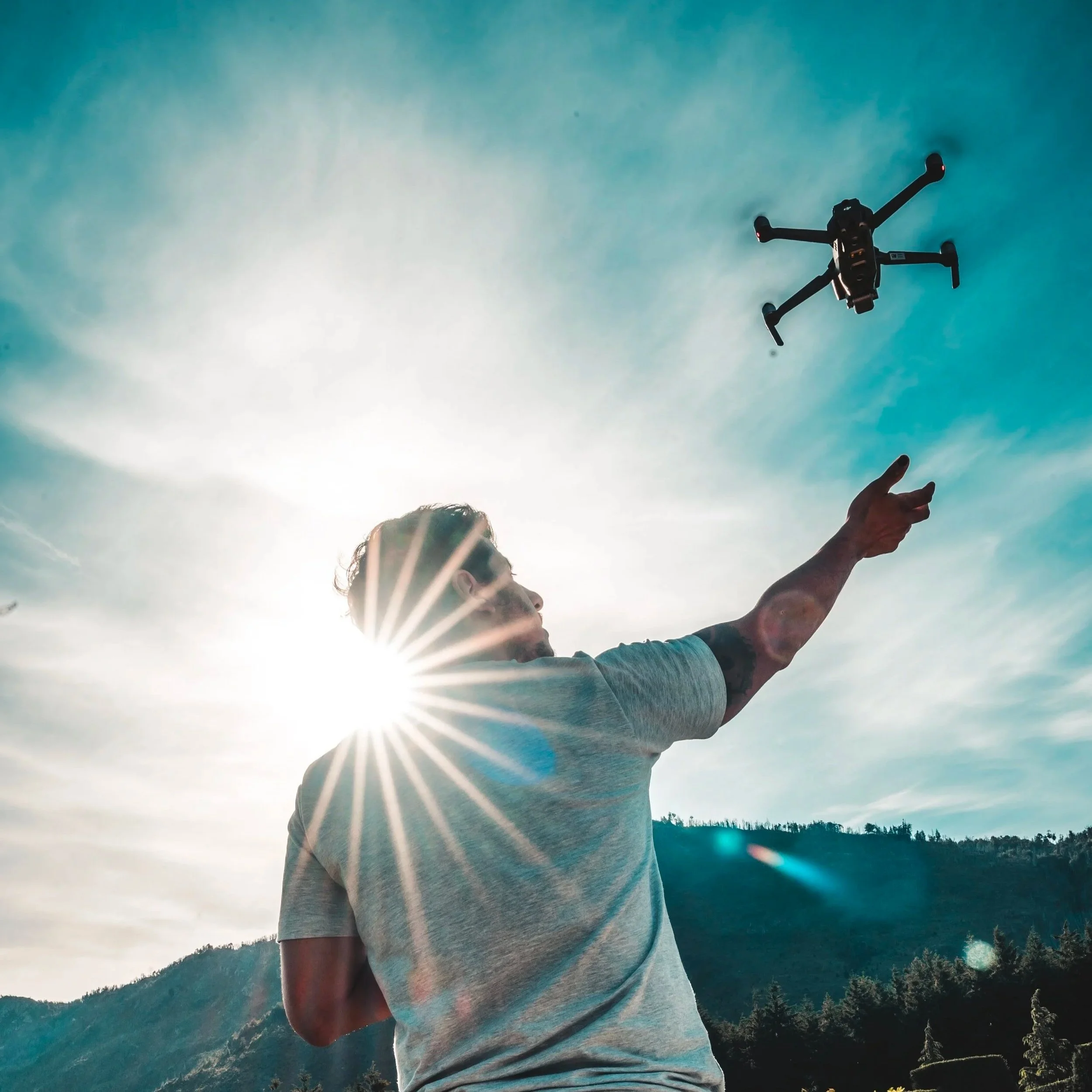 Persona realizando trucos con un dron en un paisaje montañoso bajo un cielo despejado con el sol brillante.